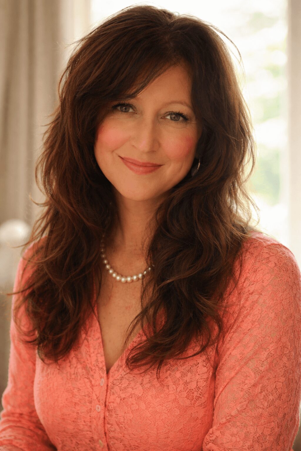 Woman with long brown hair smiling, wearing a pink top and pearl necklace, indoors with a window in background.