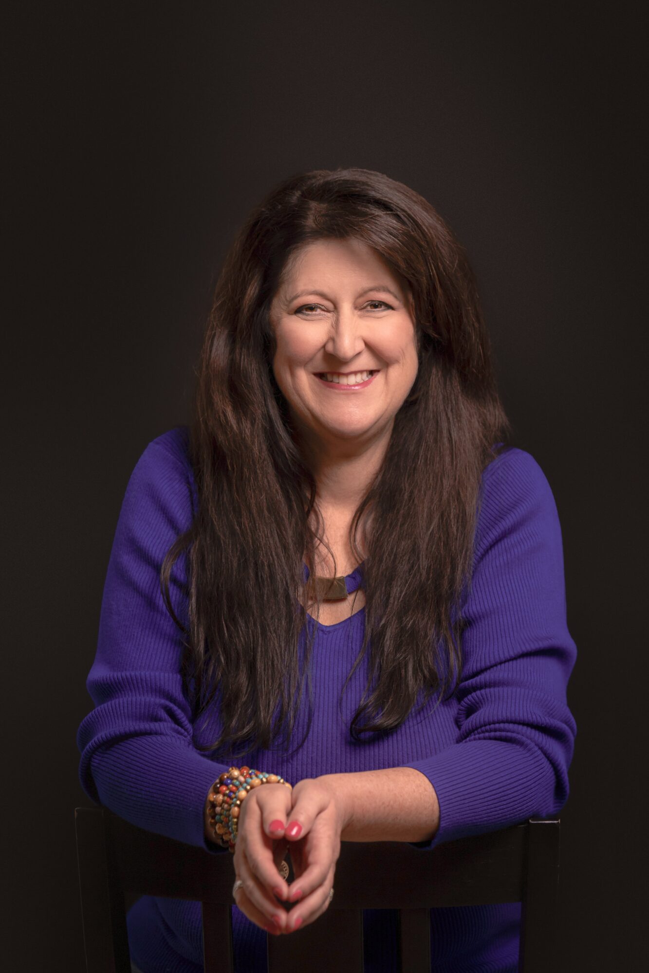 Smiling woman with long dark hair wearing a purple top, sitting with hands clasped, dark background.