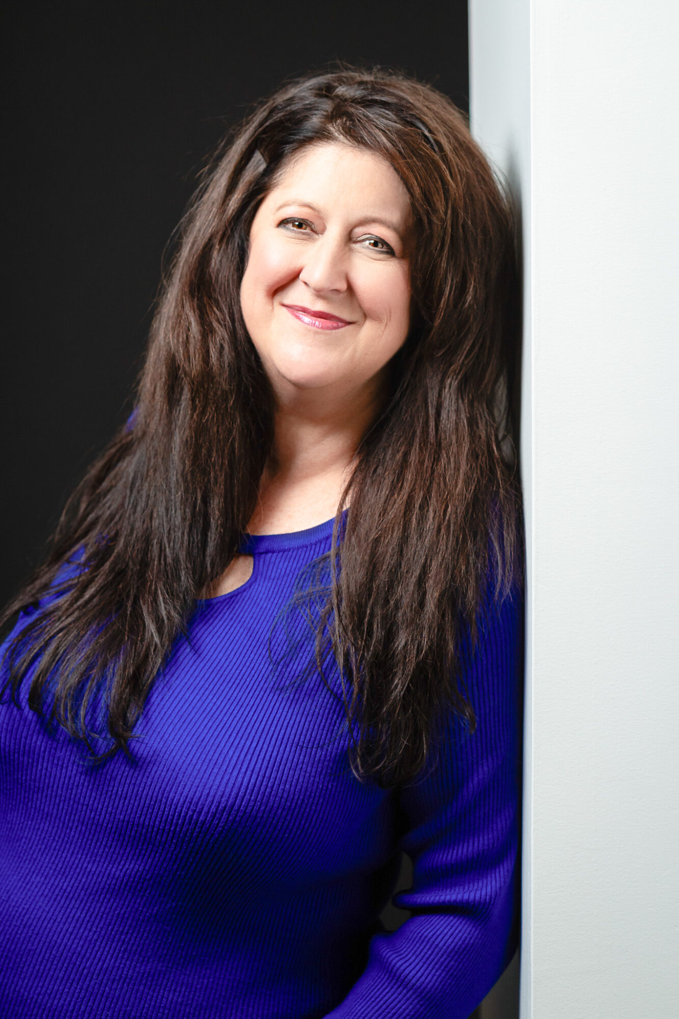 Woman with long dark hair smiling, wearing a blue top, standing against a black and white background.