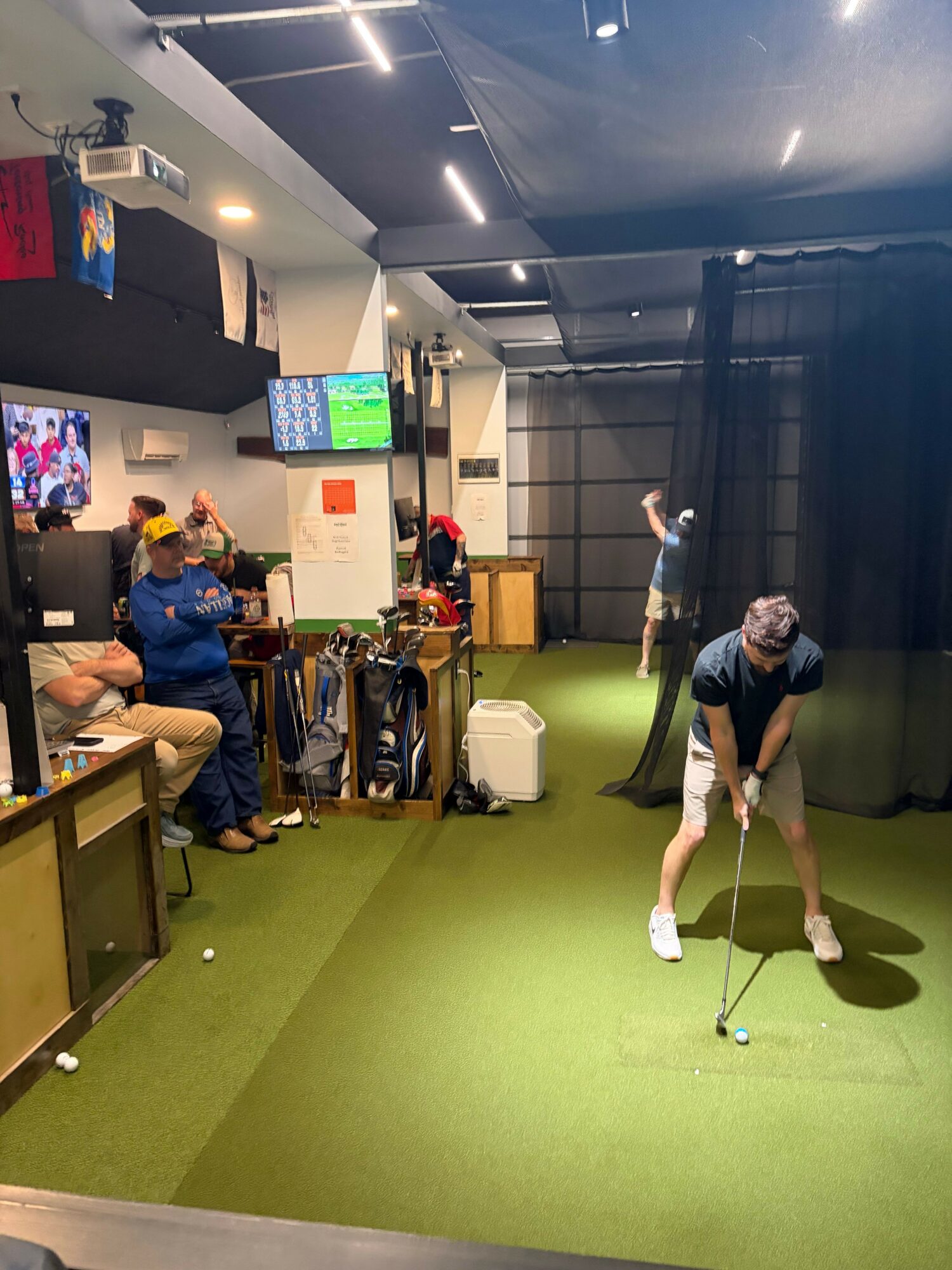 Indoor golf simulation room with a person preparing to hit a golf ball, others watching, and a screen displaying a game.