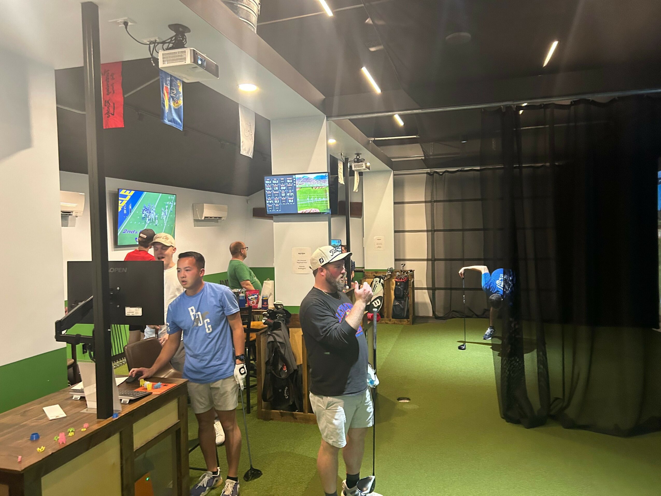 Indoor gaming or simulation room with people, screens, and a green floor, some playing or observing activities.