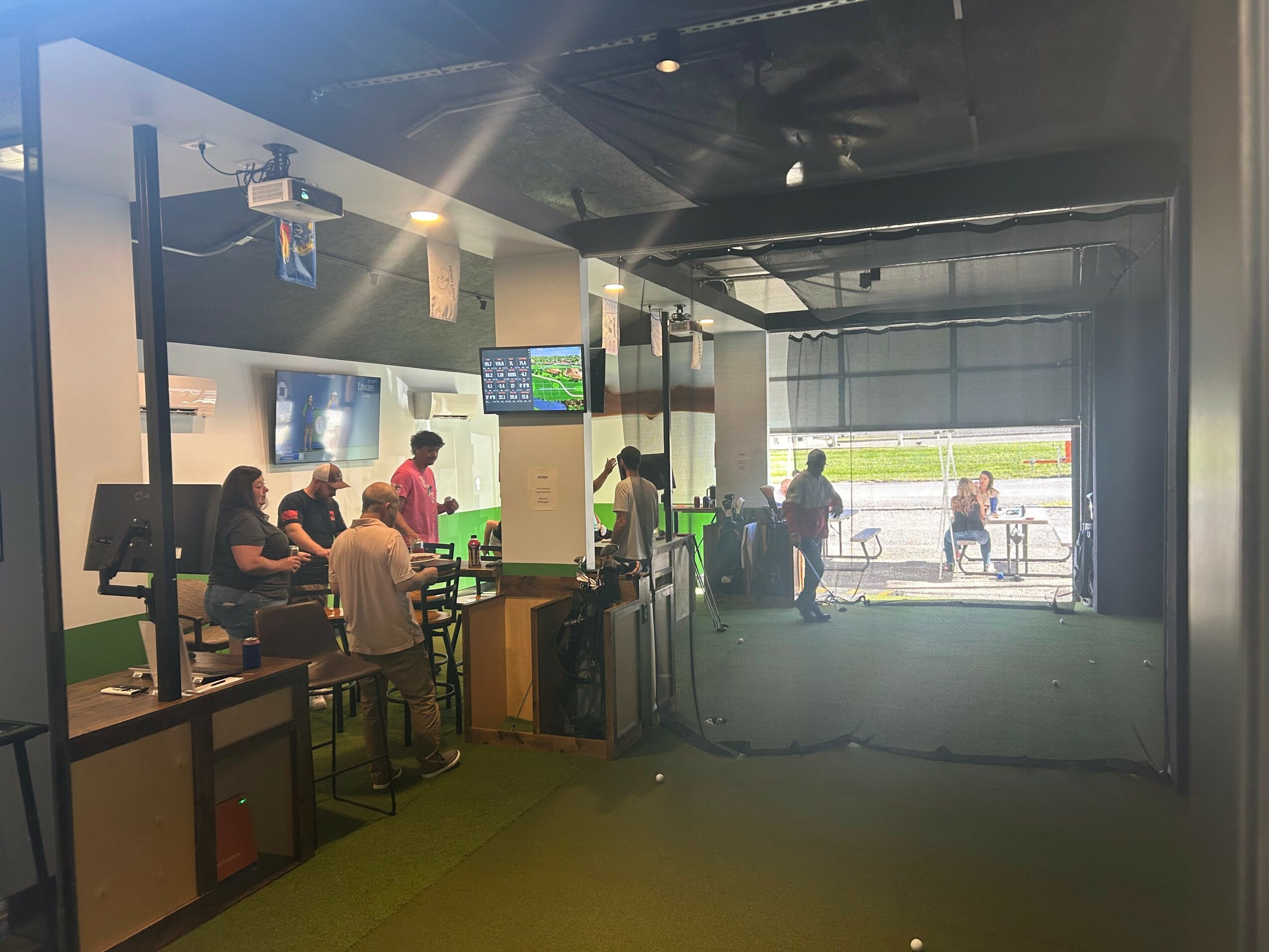 Indoor area with people sitting and standing, large windows showing outdoor scene, multiple screens on walls, green flooring.