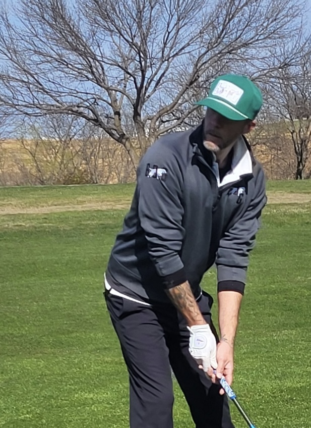 Person wearing a cap and jacket, preparing to swing a golf club on a grassy course with trees in background.