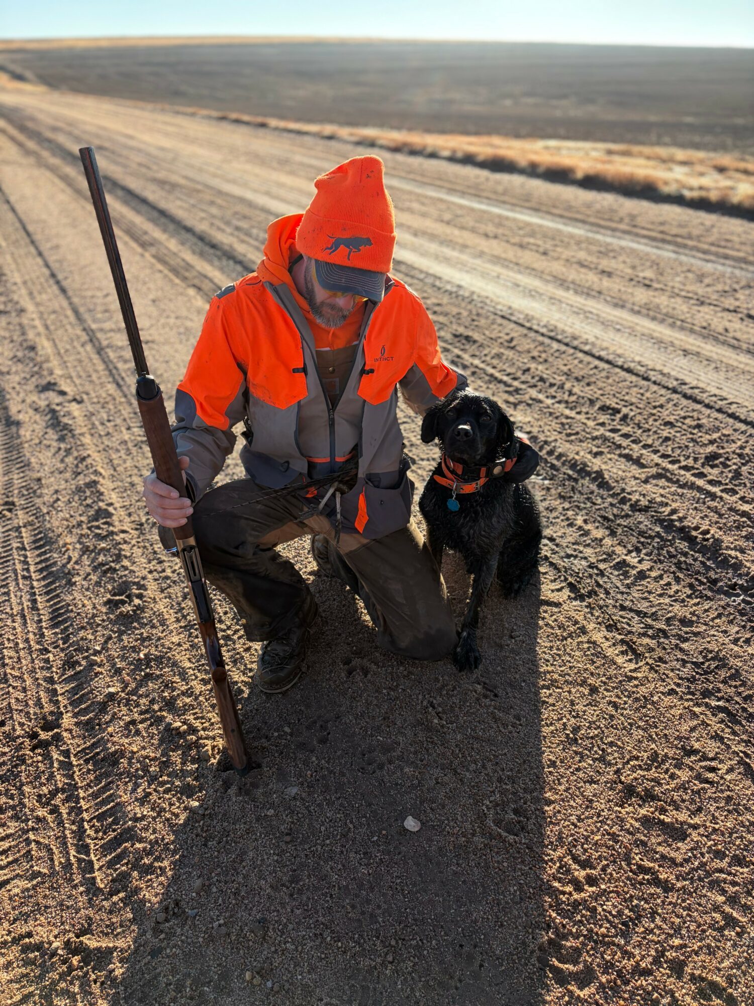 Person in orange jacket and hat kneeling beside black dog on dirt road, holding a walking stick, in open landscape.