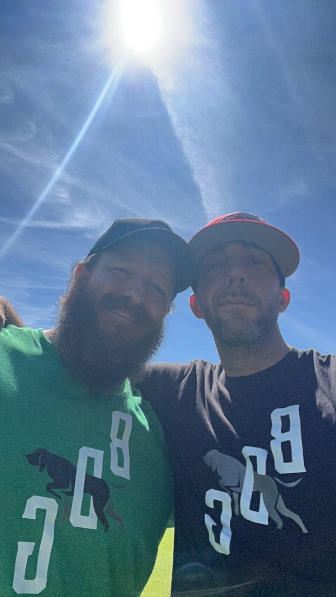 Two people taking a selfie outdoors under a bright sun with a blue sky and clouds.