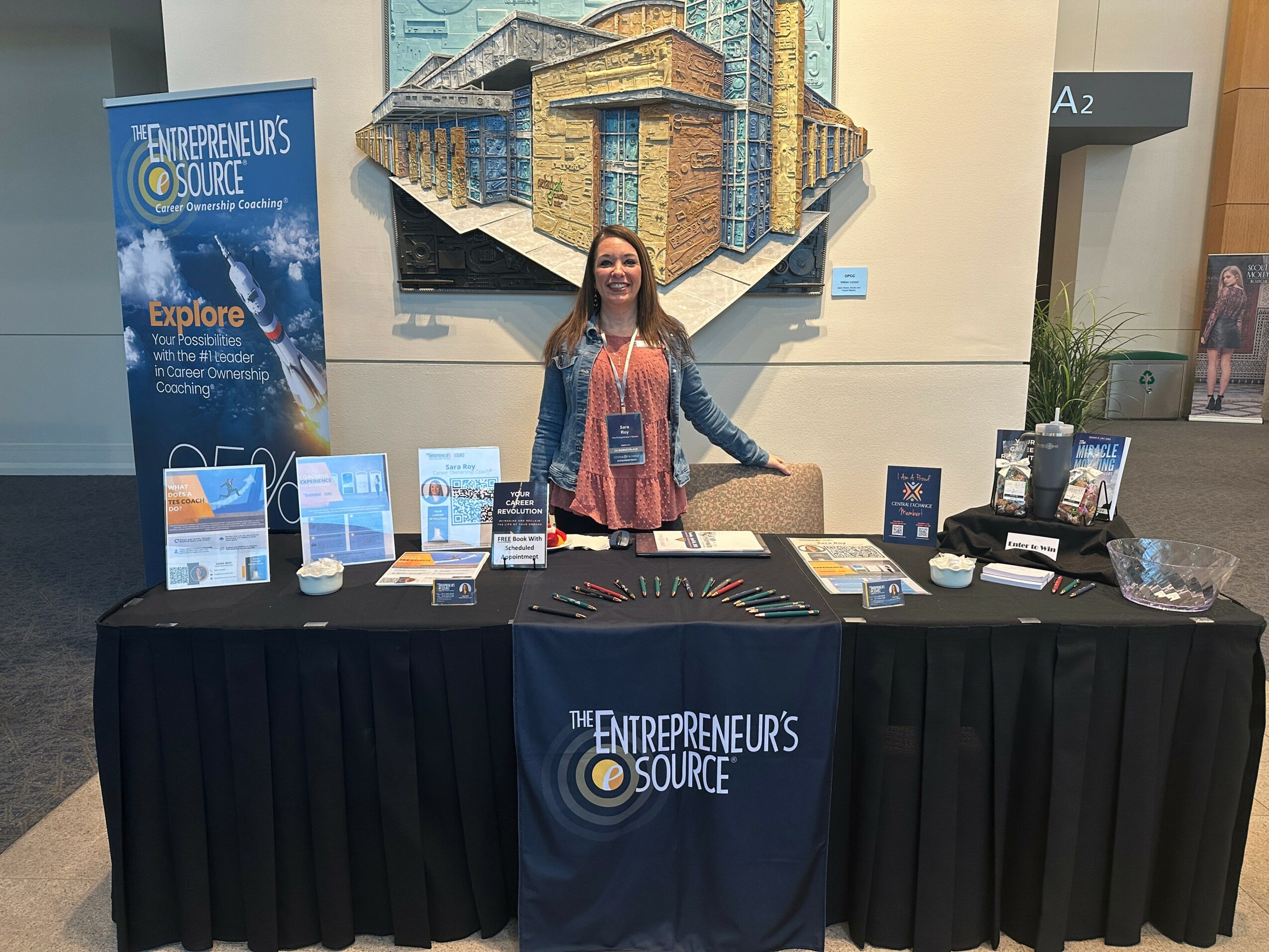 Woman standing behind a table with promotional materials at an event booth, with a large colorful artwork behind her.