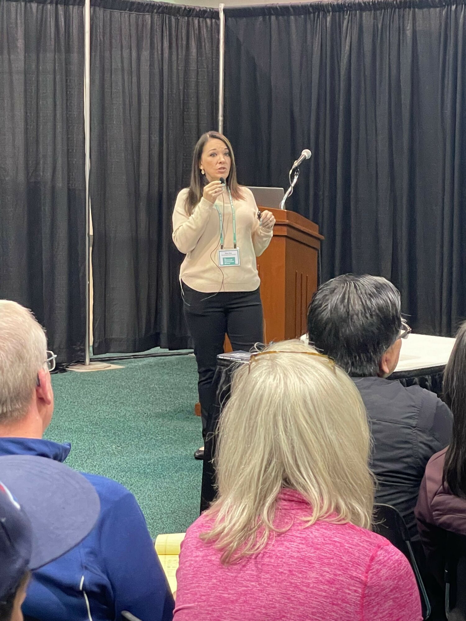 Woman speaking at podium in front of black curtains, audience seated, some taking notes, conference setting.