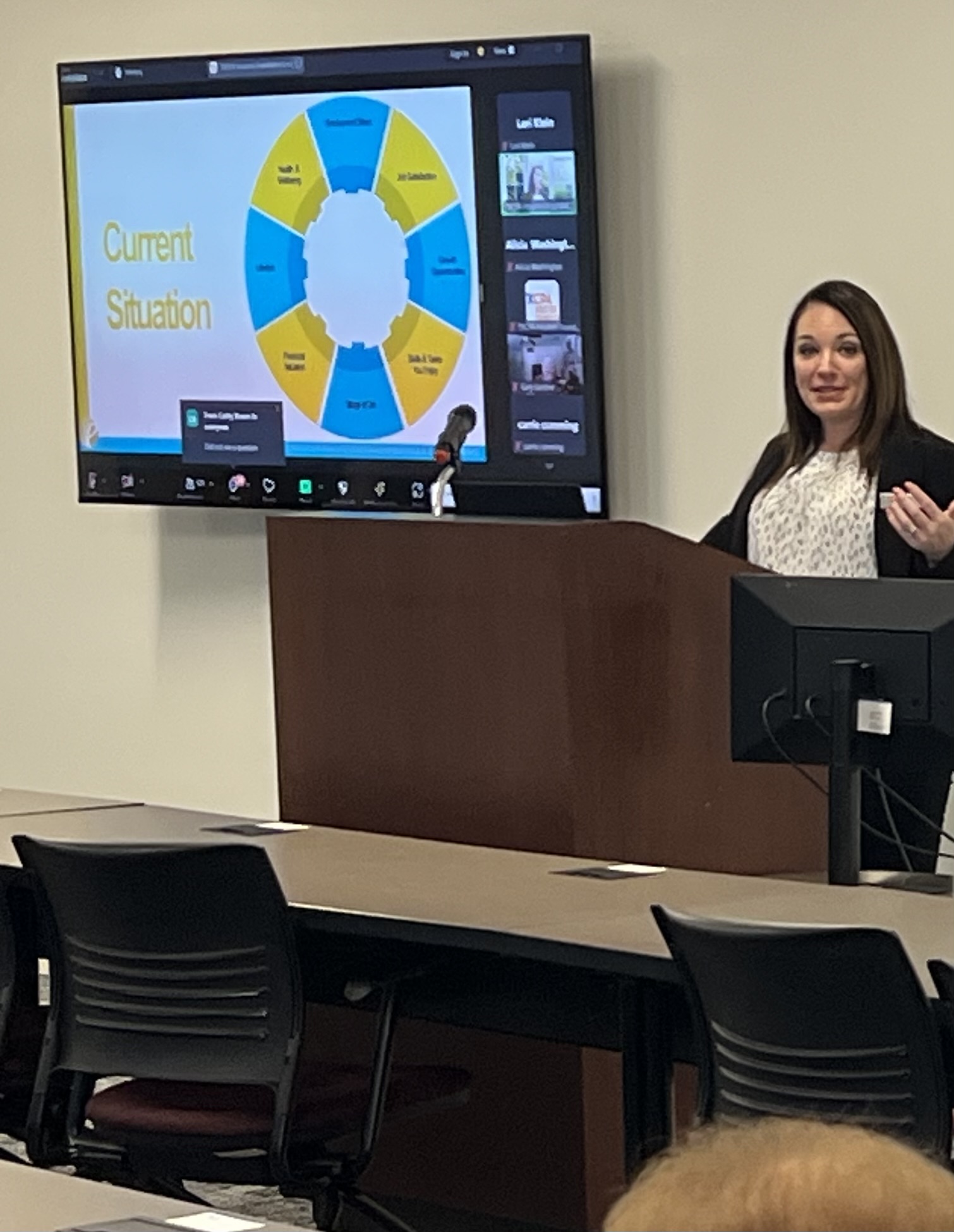 Woman presenting in front of a large screen displaying a diagram with yellow and blue sections, in a classroom or conference room.