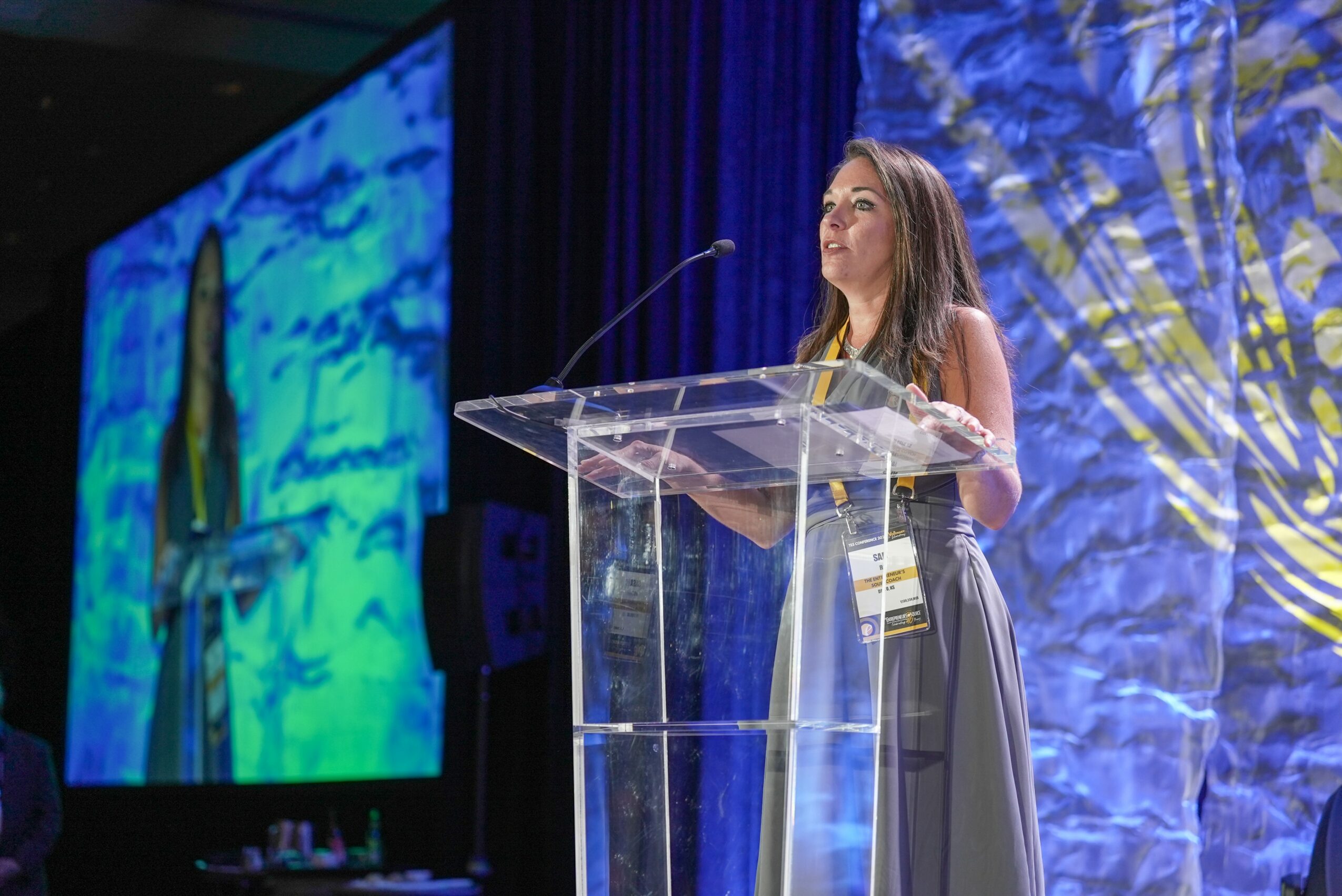 Woman standing at a clear podium speaking at a conference with large colorful screens behind her.
