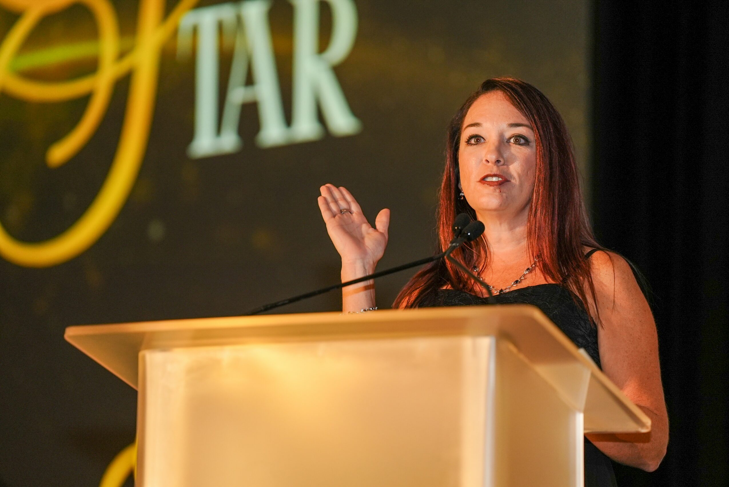 Woman speaking at a podium with a large screen behind her displaying text and graphics.