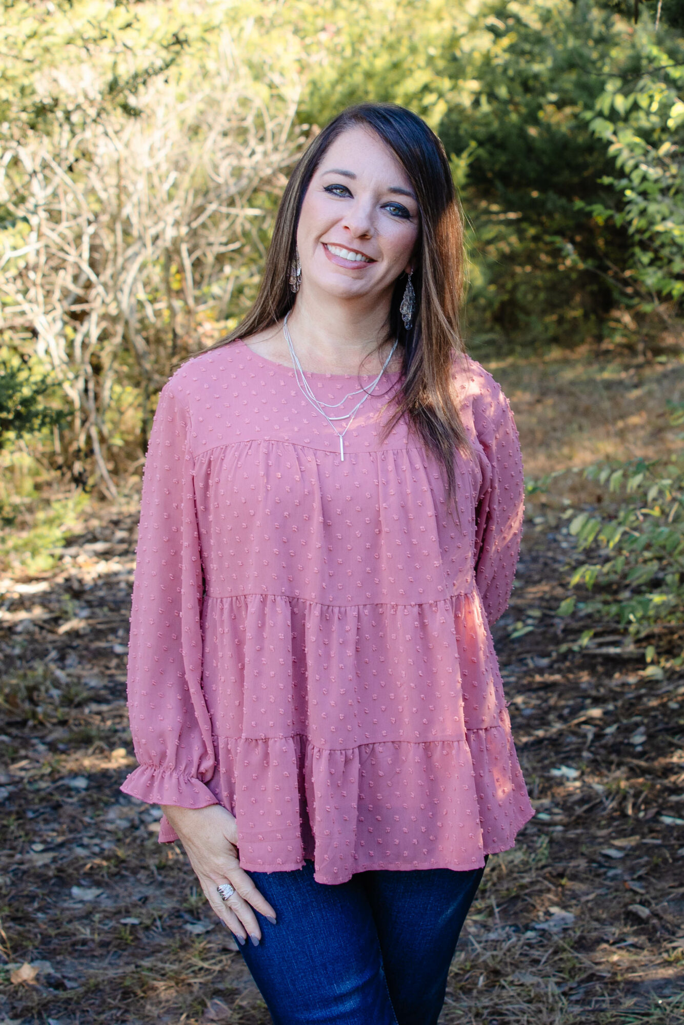 Woman with long dark hair smiling outdoors, wearing a pink long-sleeve top and blue jeans.