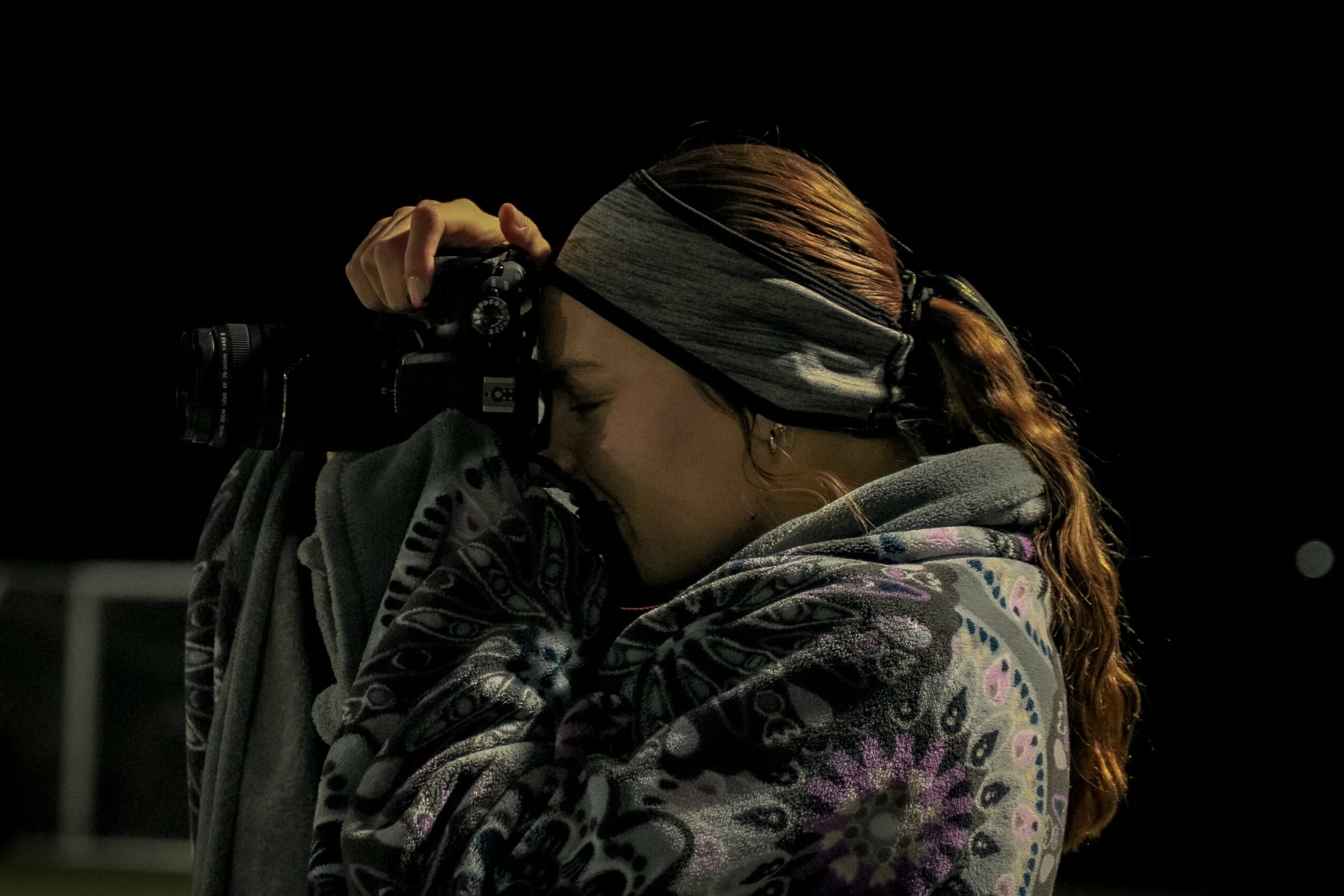 Person with long hair and headband taking a photo with a camera, wearing a patterned hoodie, against a dark background.