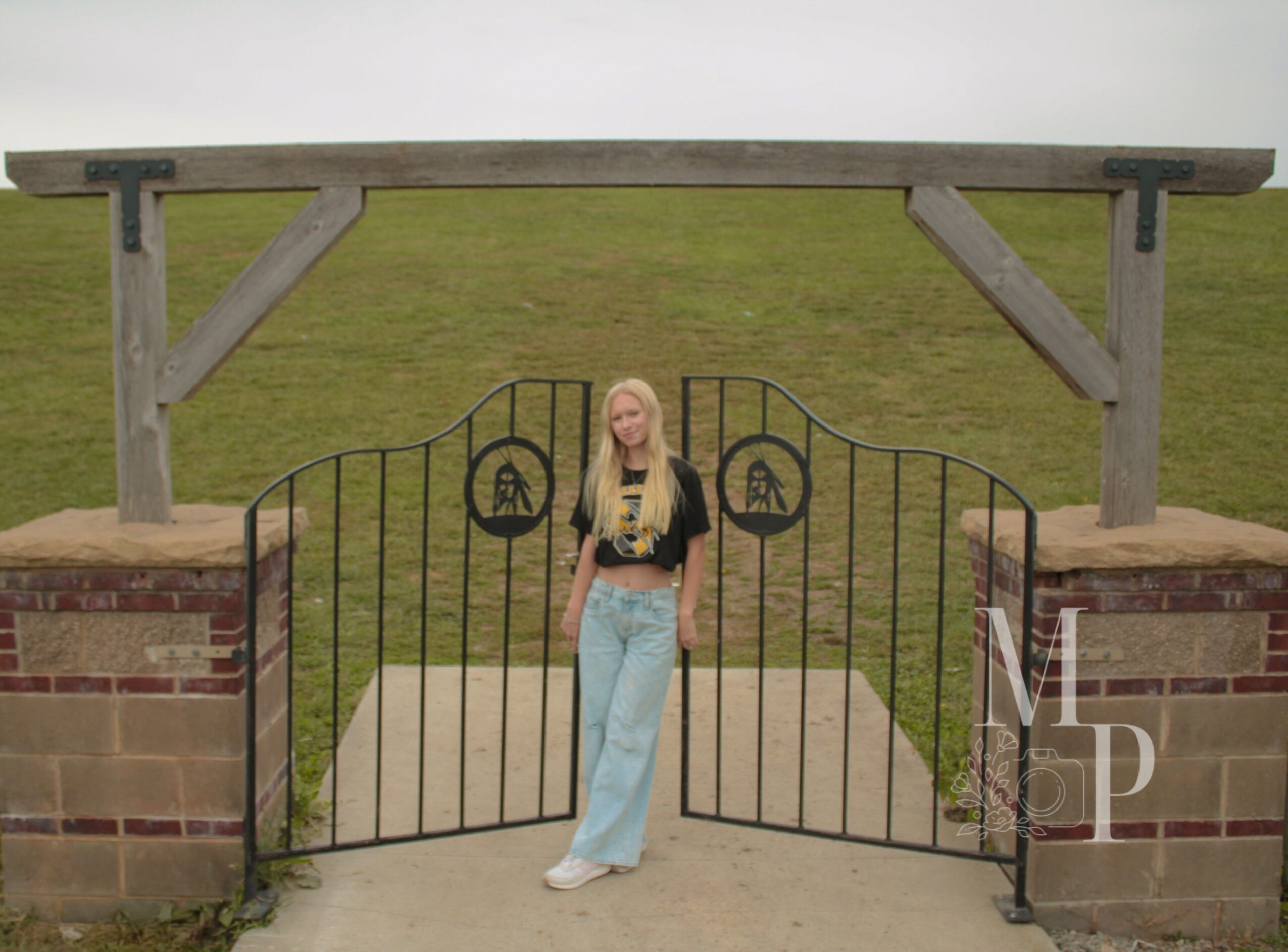 Young woman with long blonde hair standing in front of a black gate with decorative symbols, under a wooden archway.