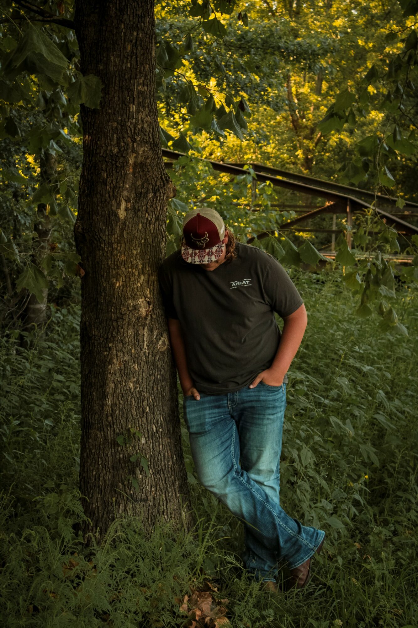 Person leaning against a tree with hands in pockets, surrounded by green foliage and yellow flowers.