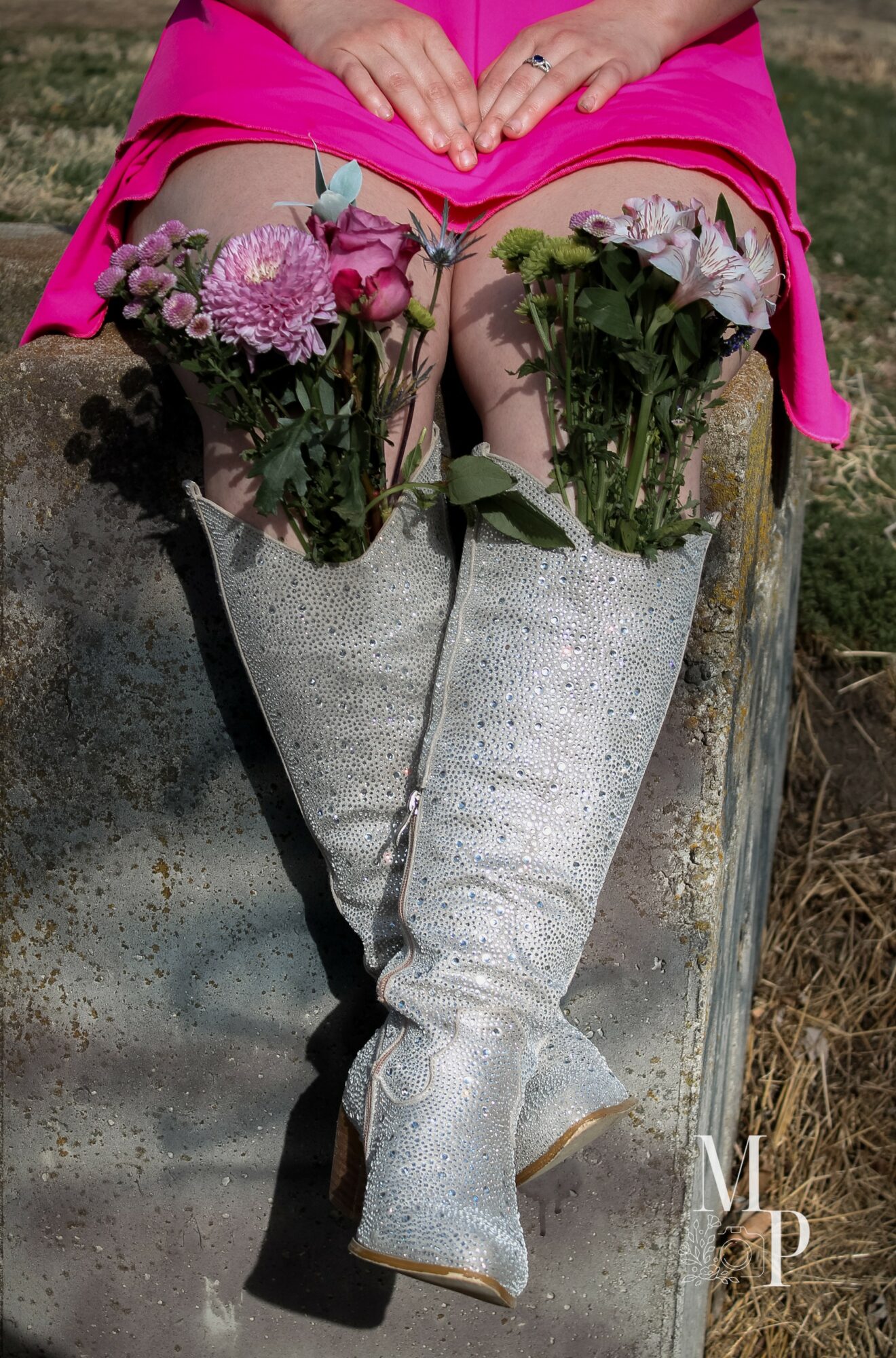 Person sitting on a metal surface with legs crossed, wearing white boots and a pink dress, holding flowers.