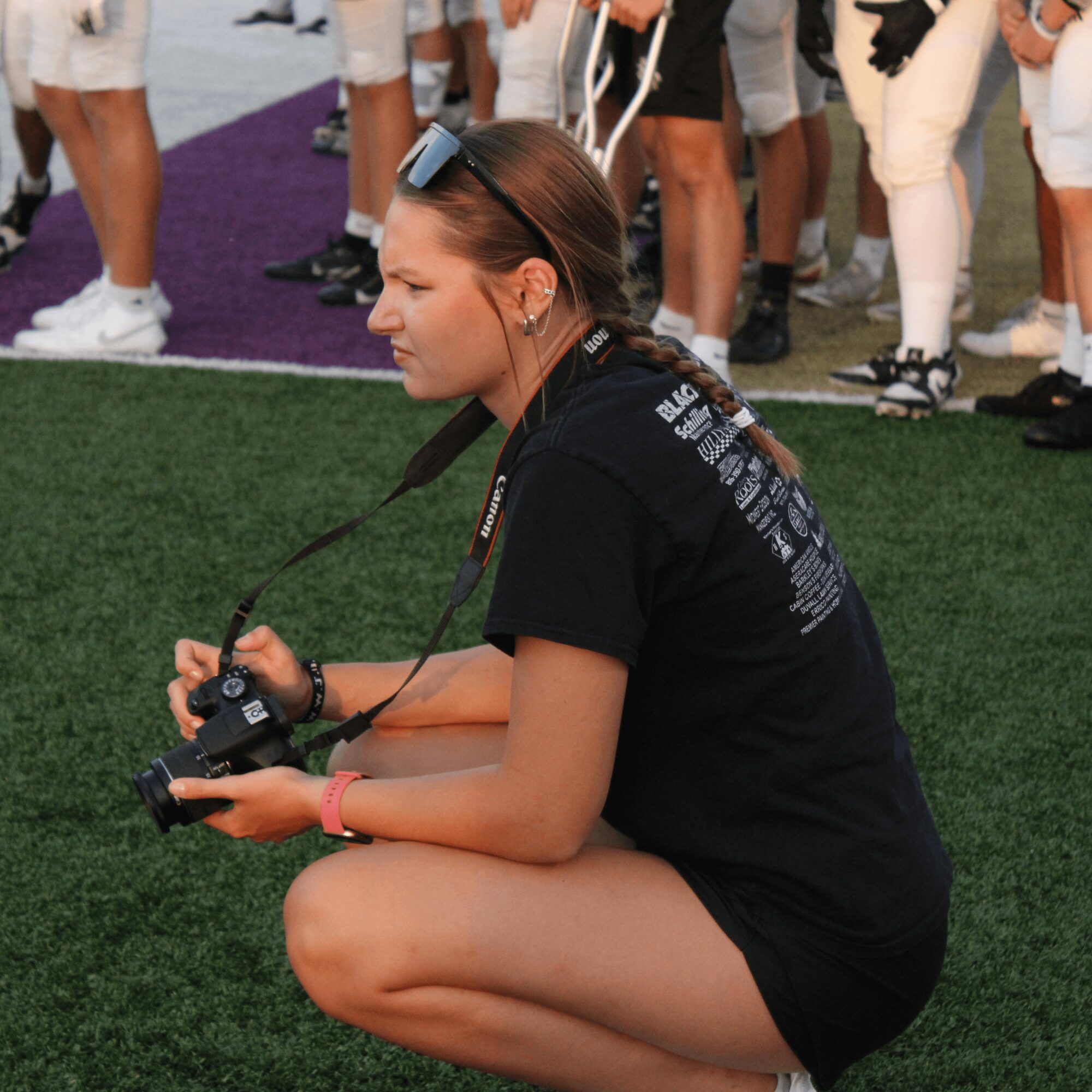 Young woman crouching on grass holding a camera, surrounded by people standing on a field.