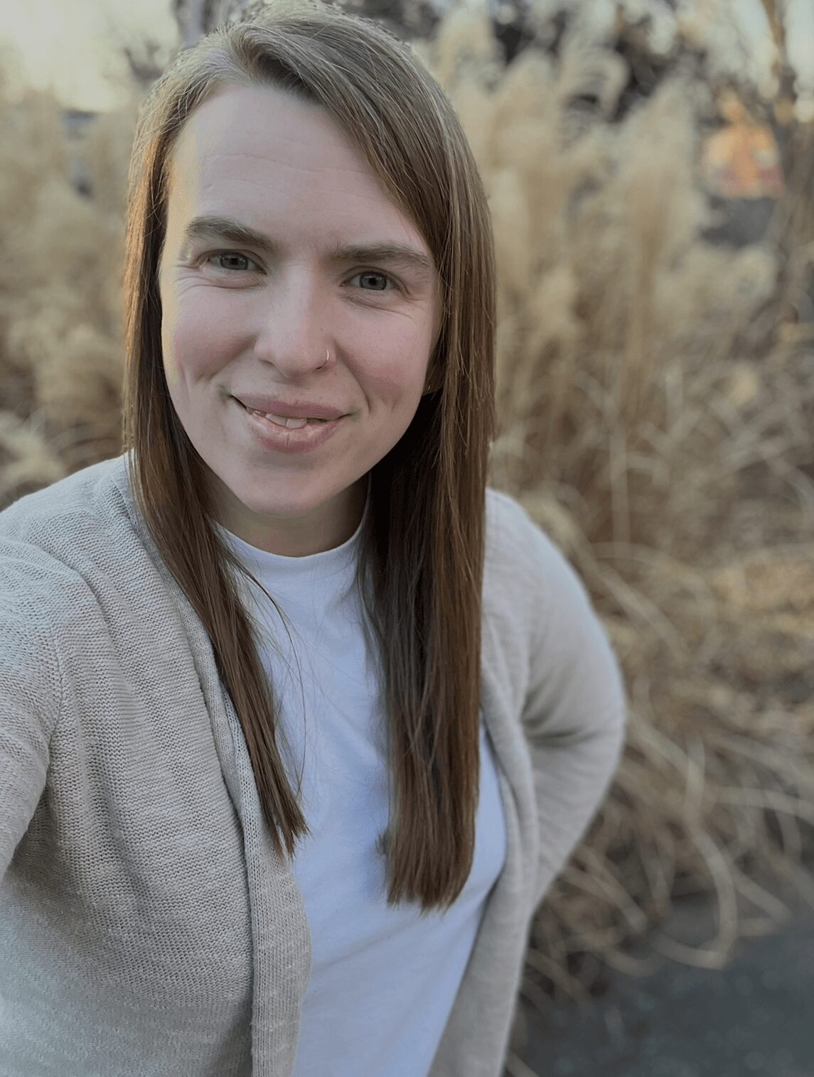 Young woman with long brown hair smiling outdoors with blurred plants in background.