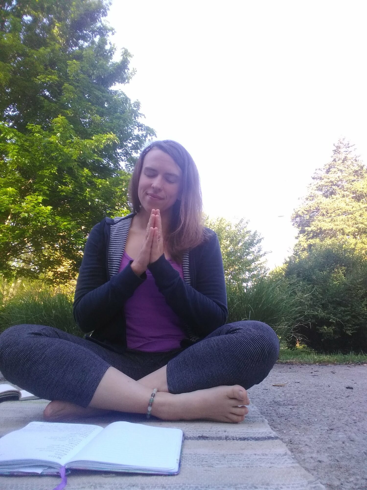 Young woman sitting cross-legged outdoors with hands in prayer position, eyes closed, surrounded by trees and greenery.