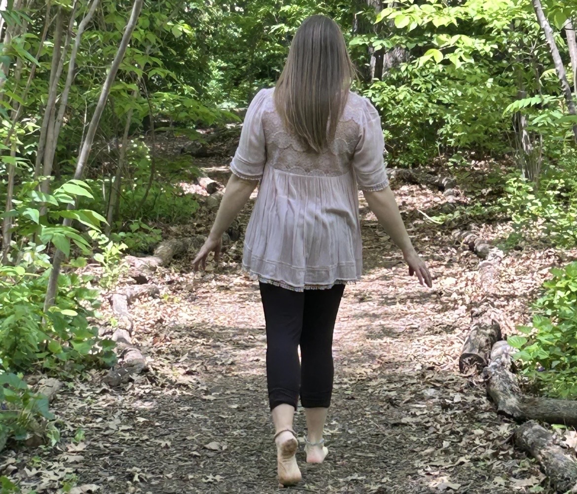 Person with long hair walking on a forest trail surrounded by green trees and plants.