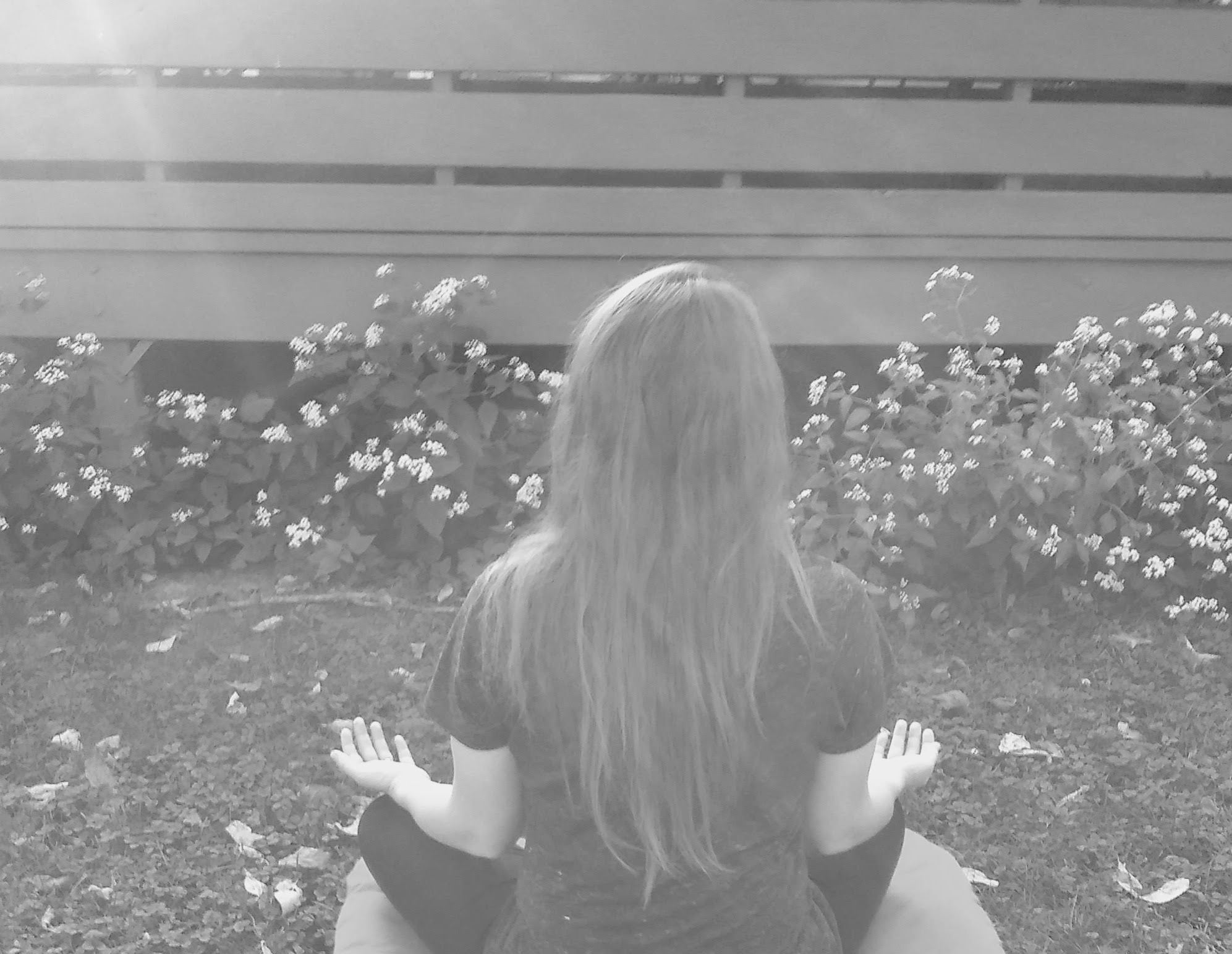 Person with long hair sitting outdoors facing a flower bed and a fence.