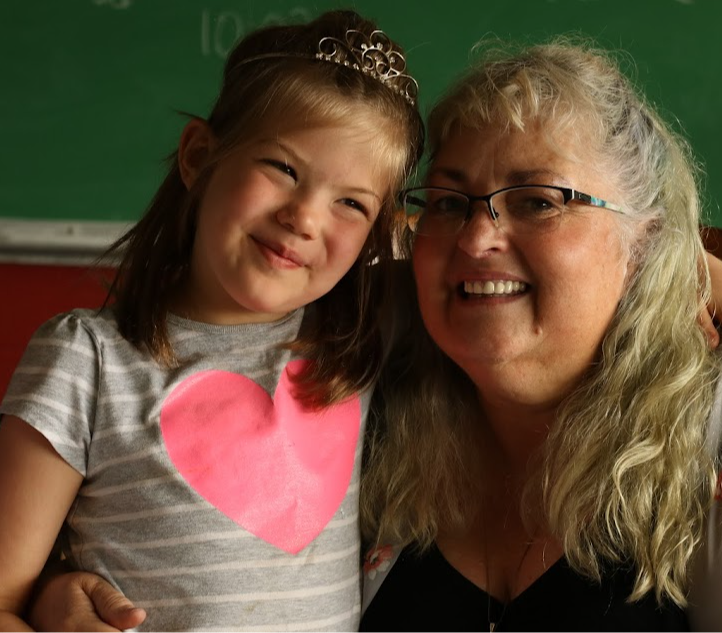 Young girl and woman smiling, girl wearing a gray striped shirt with a pink heart, woman with glasses and long blonde hair.