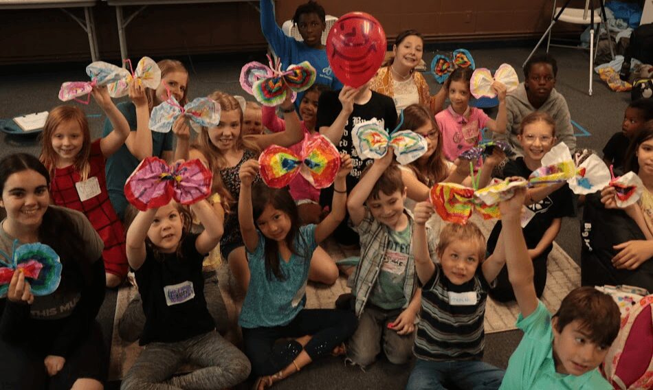 Group of children holding colorful butterfly-shaped crafts and balloons, smiling indoors.