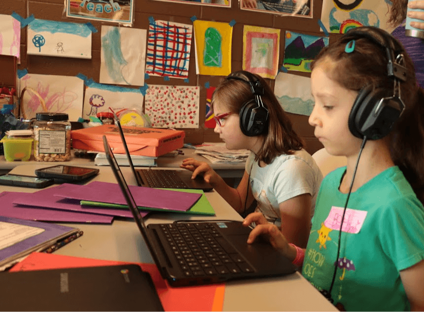 Two children wearing headphones work on laptops at a colorful table with artwork on the wall behind them.