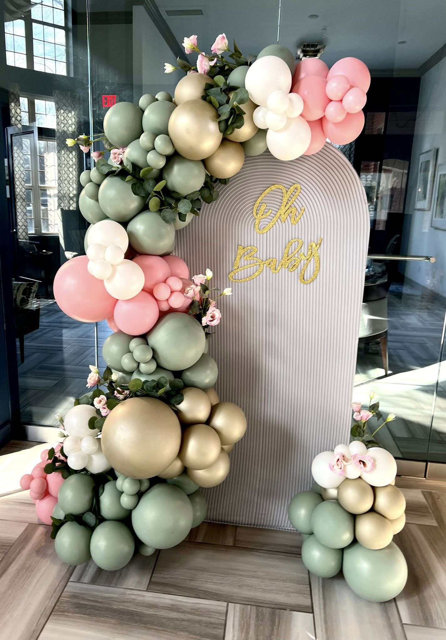 Decorative balloon arrangement with pastel colors and flowers, with a white arch and gold lettering, in a modern indoor setting.