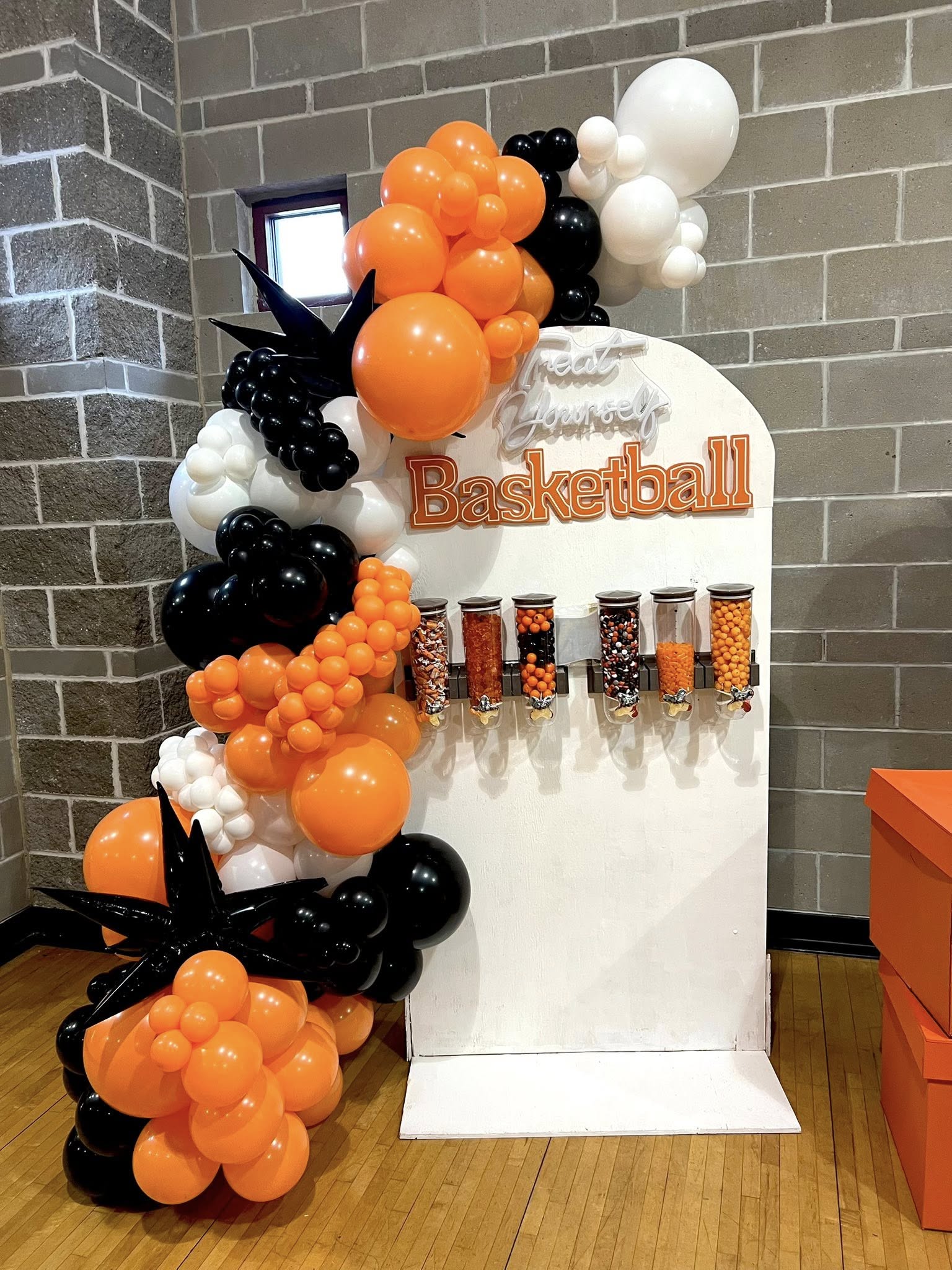 Basketball-themed display with orange, black, and white balloons arranged around a white stand against a brick wall.