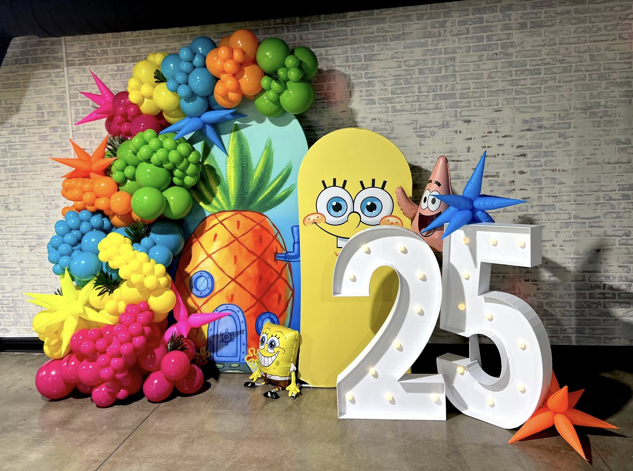 Colorful balloon display with SpongeBob character, large number 25, and pineapple decoration against a gray wall.