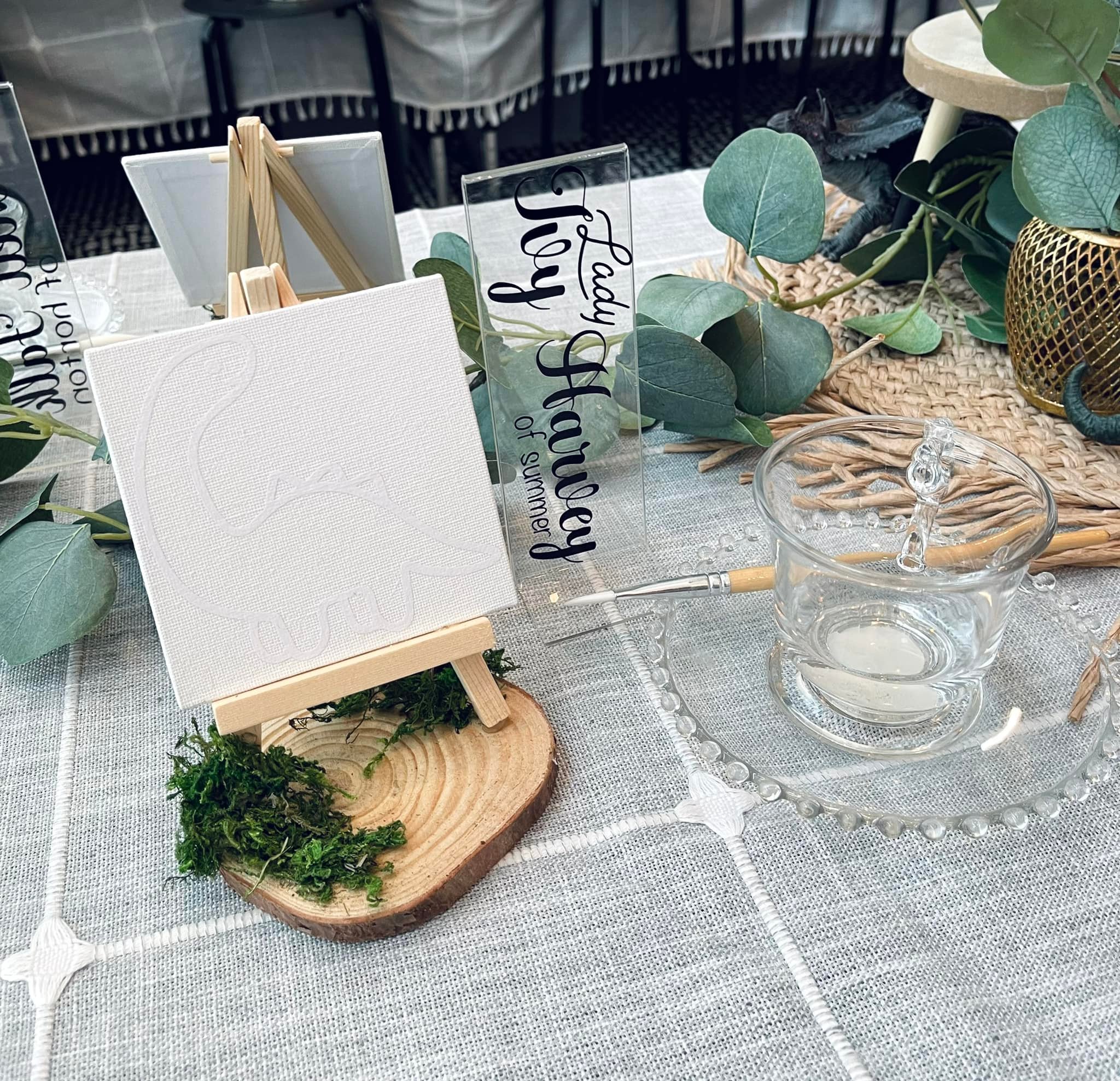 Table setting with a small easel, blank canvas, greenery, glass bowl, and decorative items on a tablecloth.