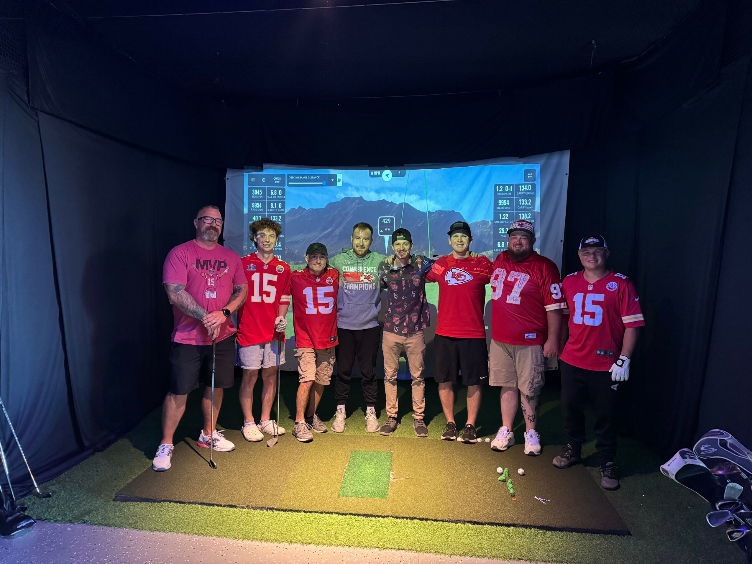 Group of seven people standing on a green mat in front of a blue screen with cityscape background, some wearing red sports jerseys.