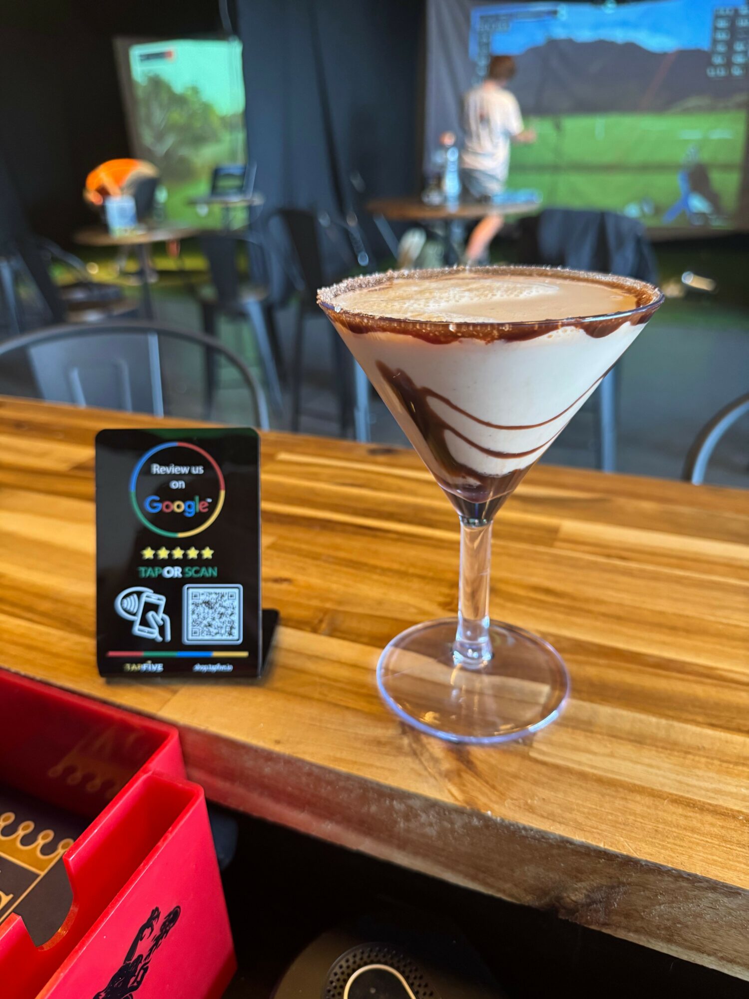 Chocolate and vanilla layered cocktail in a martini glass on a wooden bar counter, with a sports bar background.