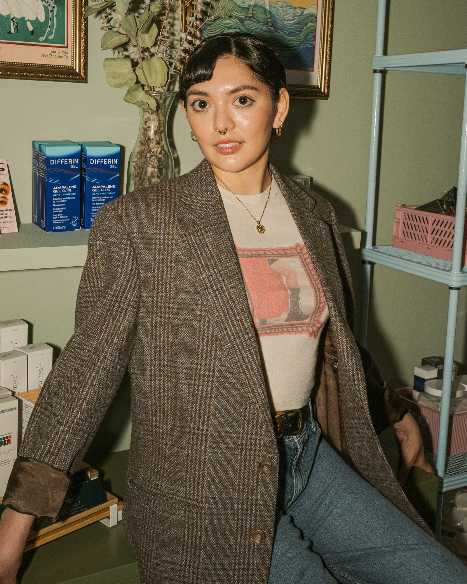 Young woman with dark hair in a bun, wearing a plaid blazer and jeans, standing indoors with shelves and paintings.
