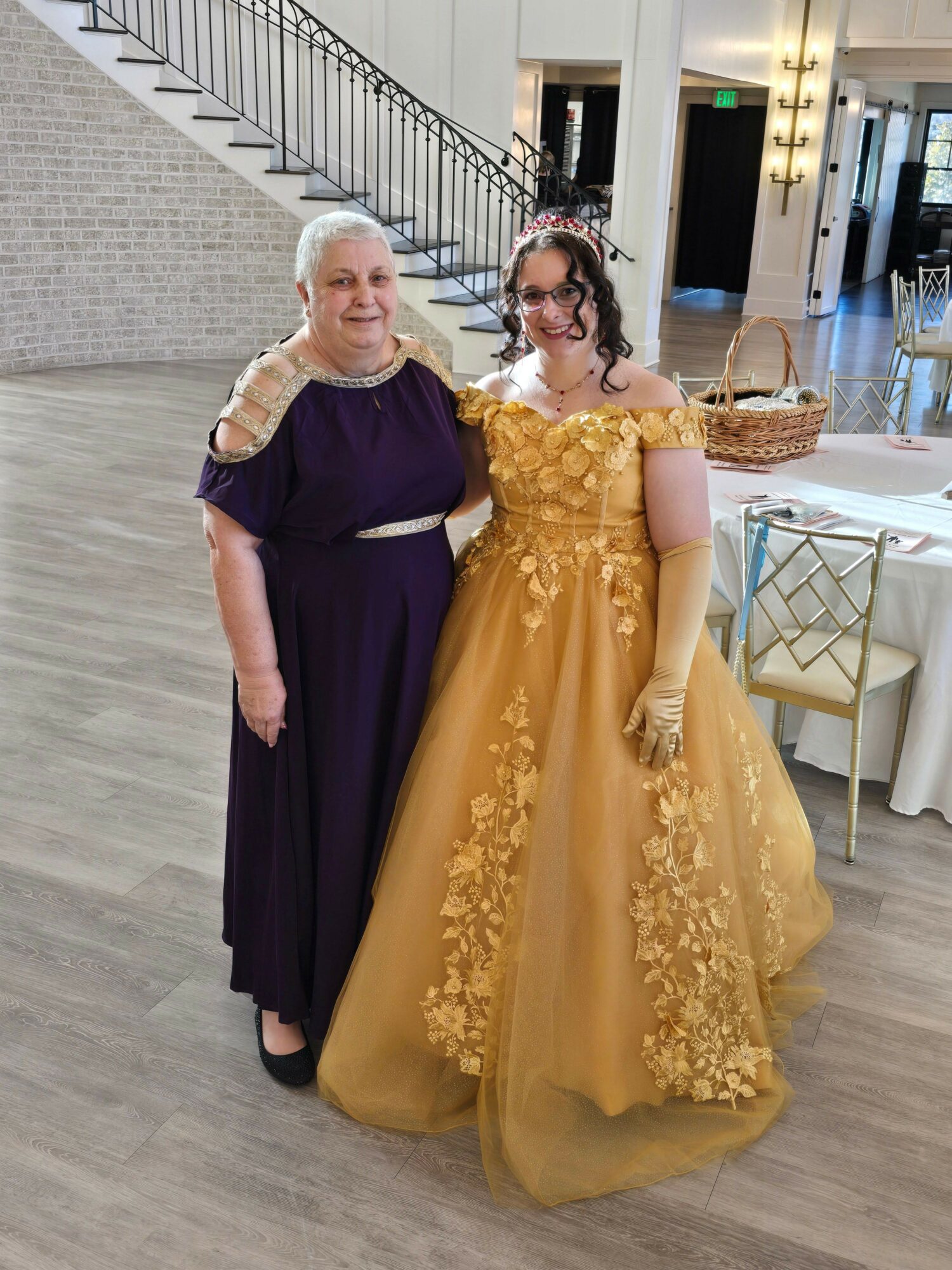 Two women in formal dresses standing indoors, one in a dark purple gown and the other in a yellow gown with floral details.