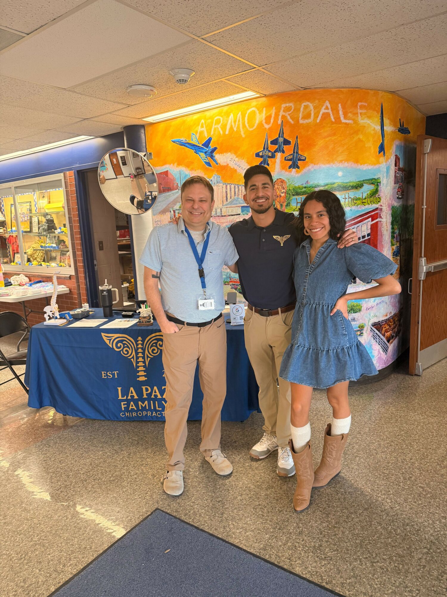 Three people standing in front of a colorful mural and a table with a blue banner, smiling at the camera.