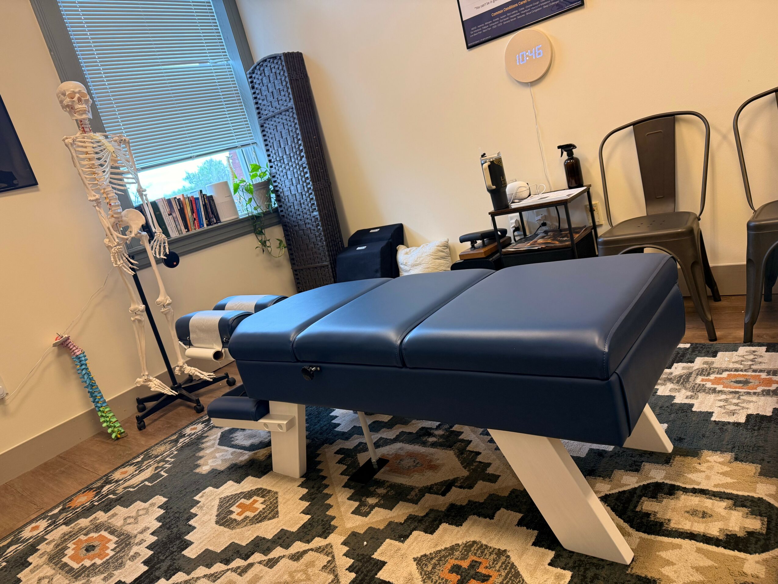 Blue examination table in a room with chairs, window, and medical equipment, on a patterned rug.