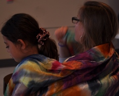 Two people sitting close, one with long hair and colorful scarf, the other with glasses and short hair.