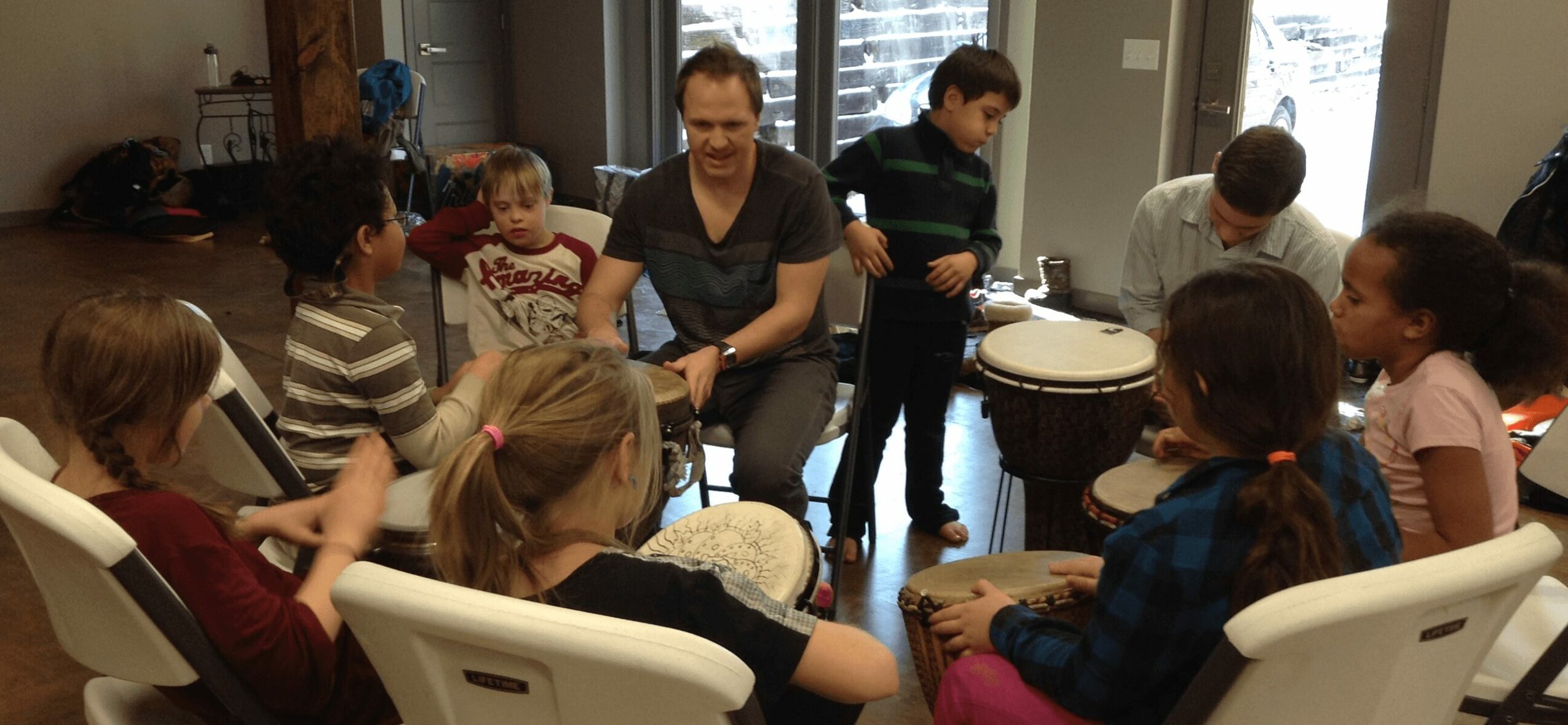 Group of children and an adult sitting in a circle indoors, some children playing drums.