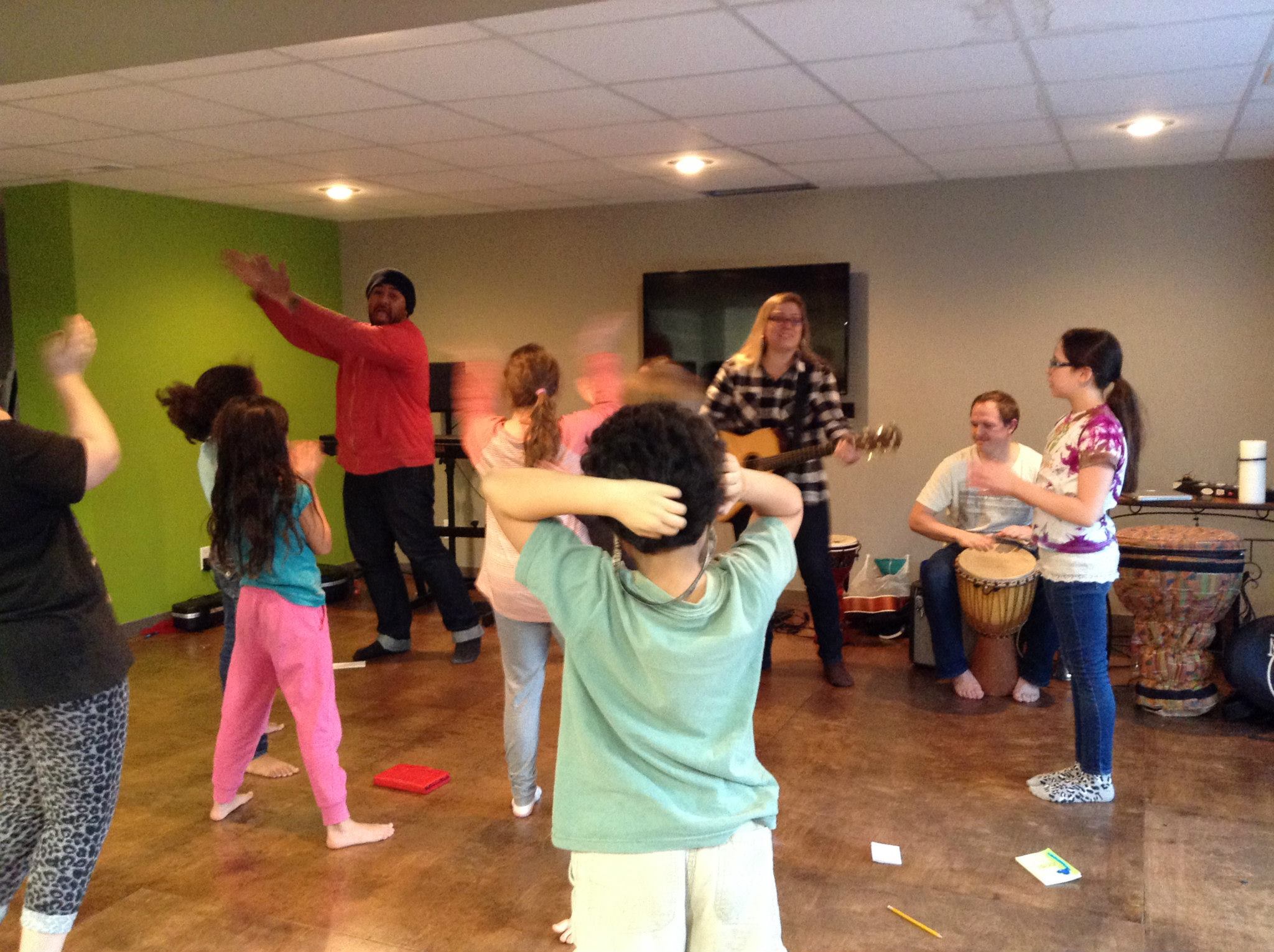 Group of people dancing and playing musical instruments in a room with green and beige walls.
