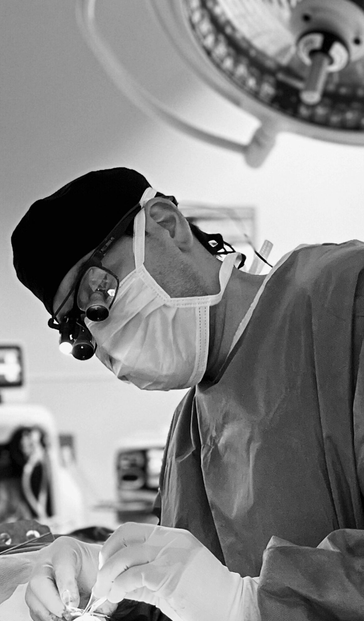 Surgeon wearing mask, glasses, and cap performing surgery in operating room with surgical light above.