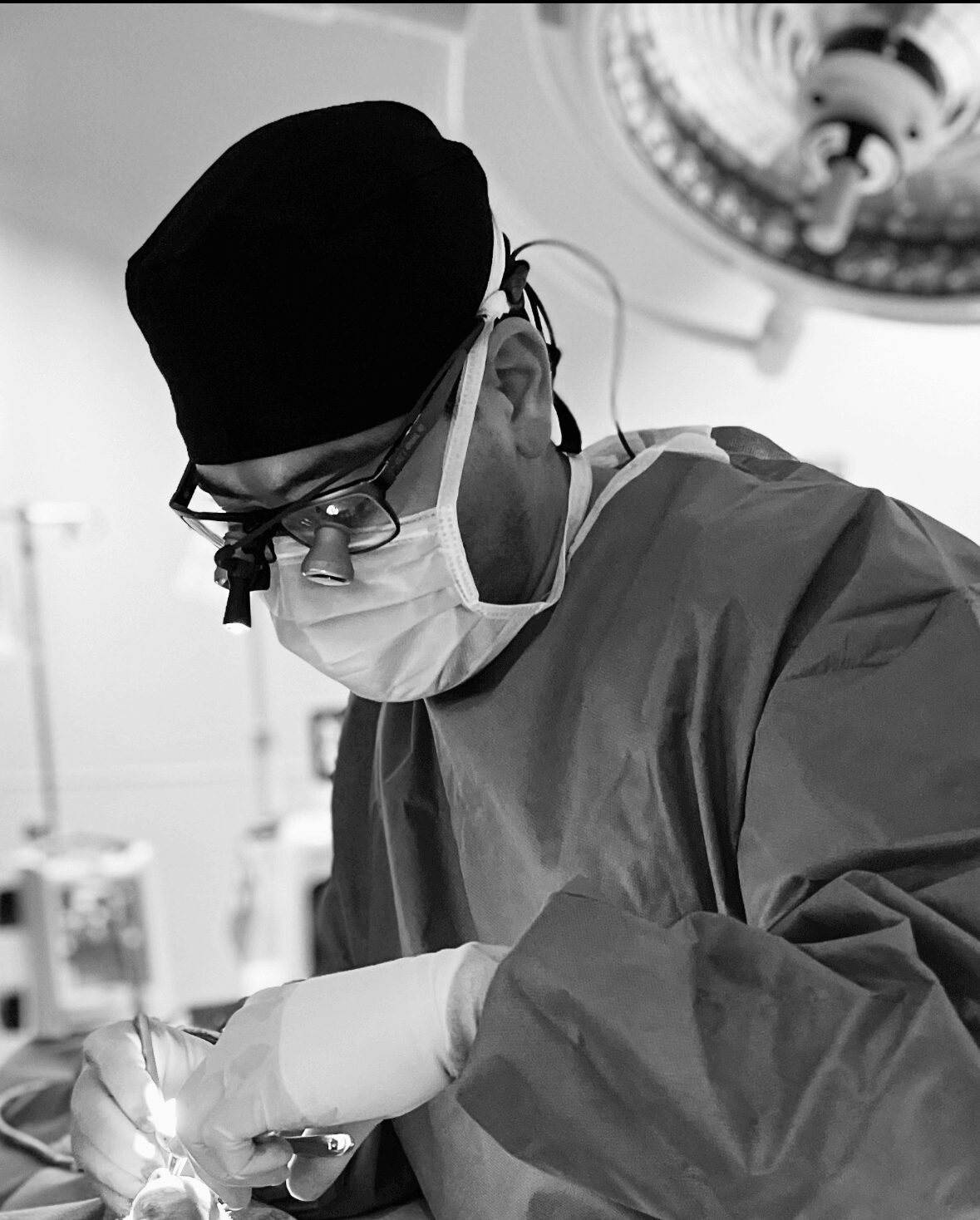 Surgeon wearing surgical mask, cap, glasses, and gown performing a procedure in an operating room.