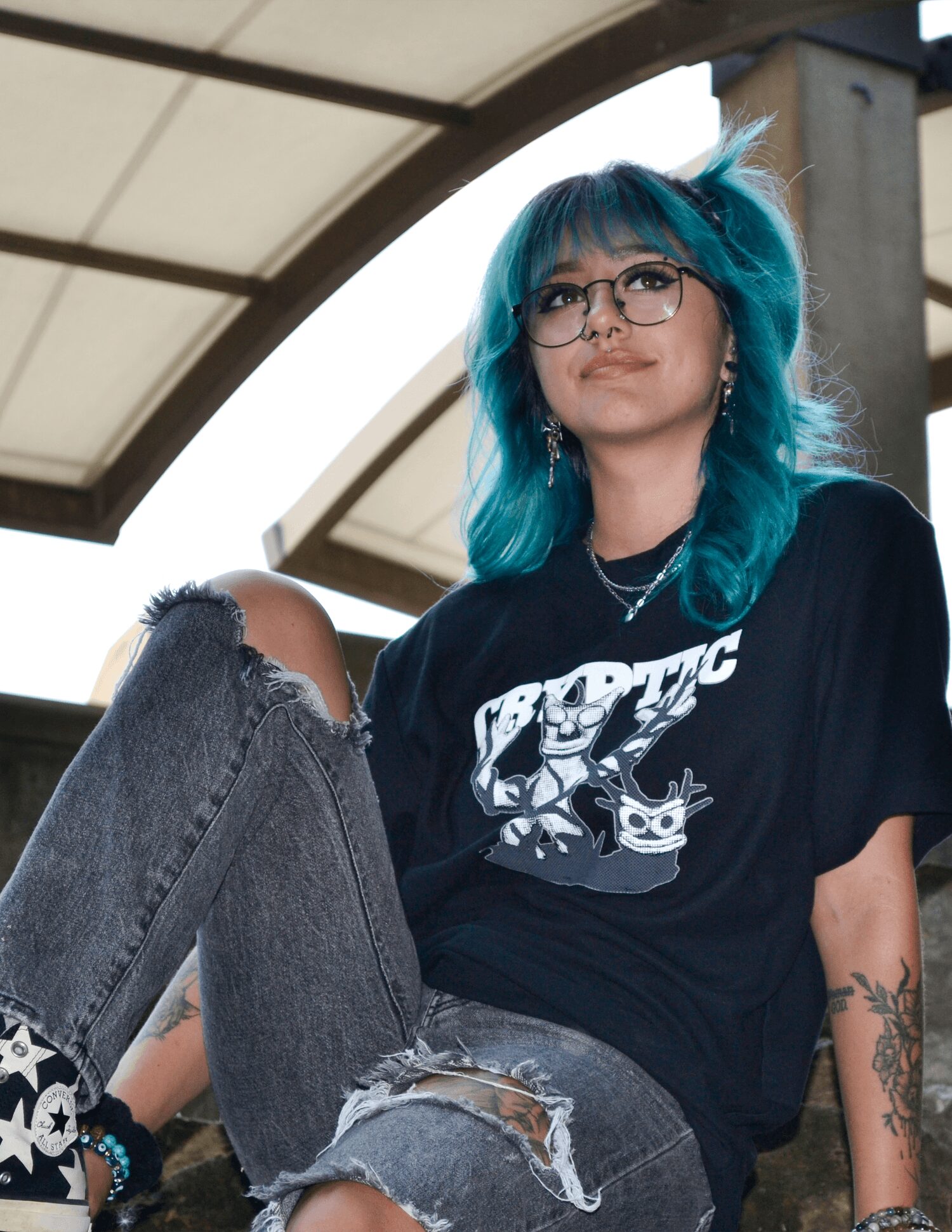 Young woman with blue hair, glasses, and tattoos, sitting outdoors with a relaxed pose, wearing a black graphic t-shirt and ripped jeans.