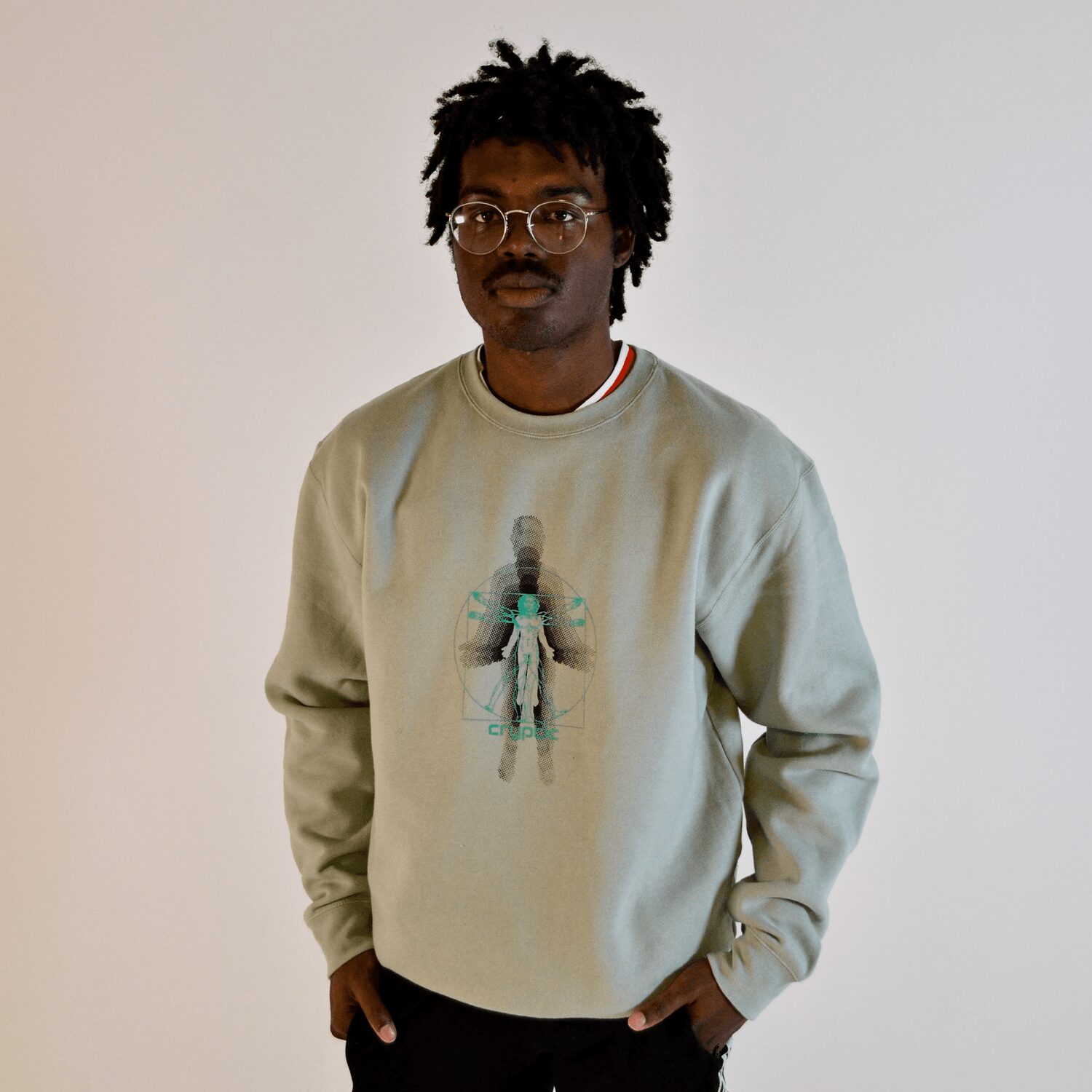 Young man with glasses and dreadlocks wearing a light sweatshirt with a graphic design, standing against a plain background.