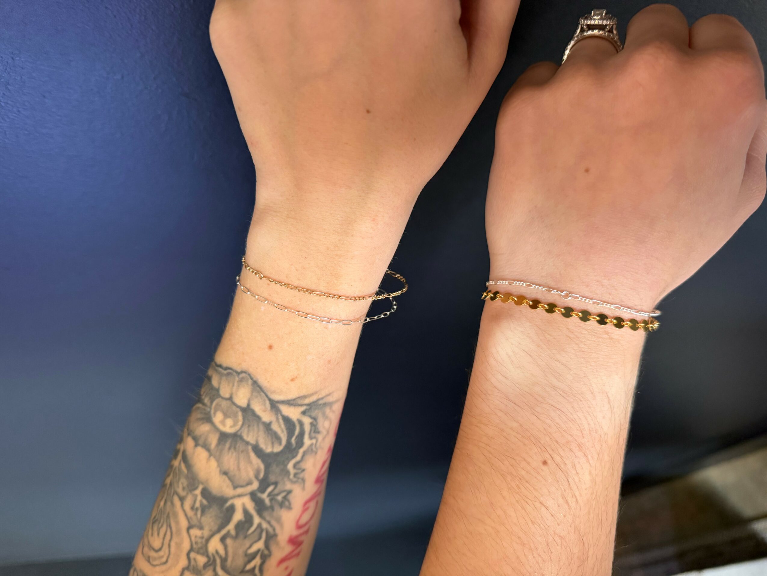 Two hands with bracelets and tattoos, one with a floral tattoo on the wrist, against a dark background.