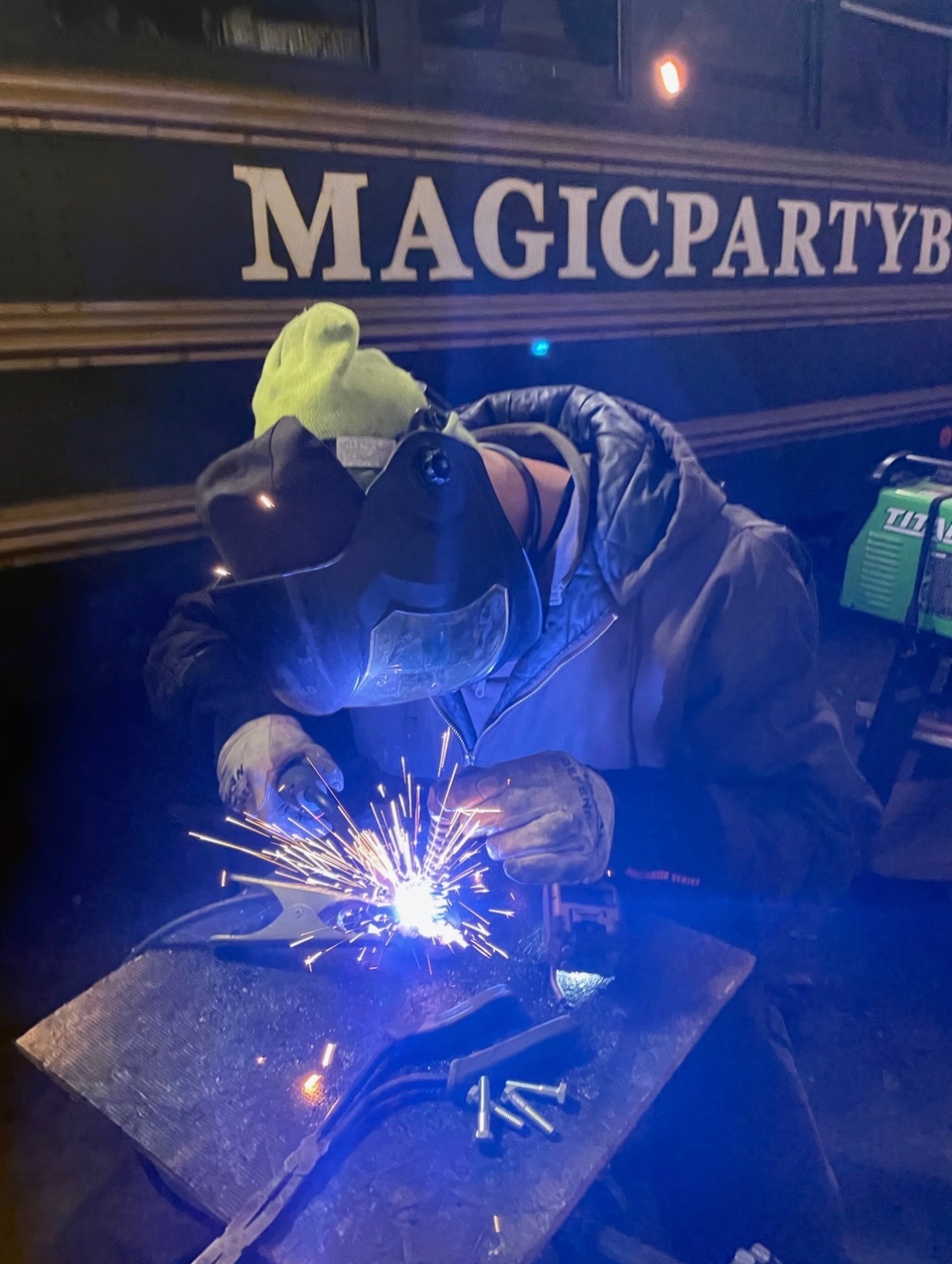 Person welding with protective gear, sparks flying, in front of a bus with 'MAGICPARTY' sign.