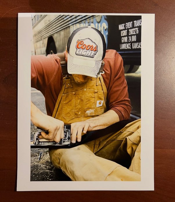 Person wearing a Coors Light cap and yellow apron, working with a tool, sitting outdoors.