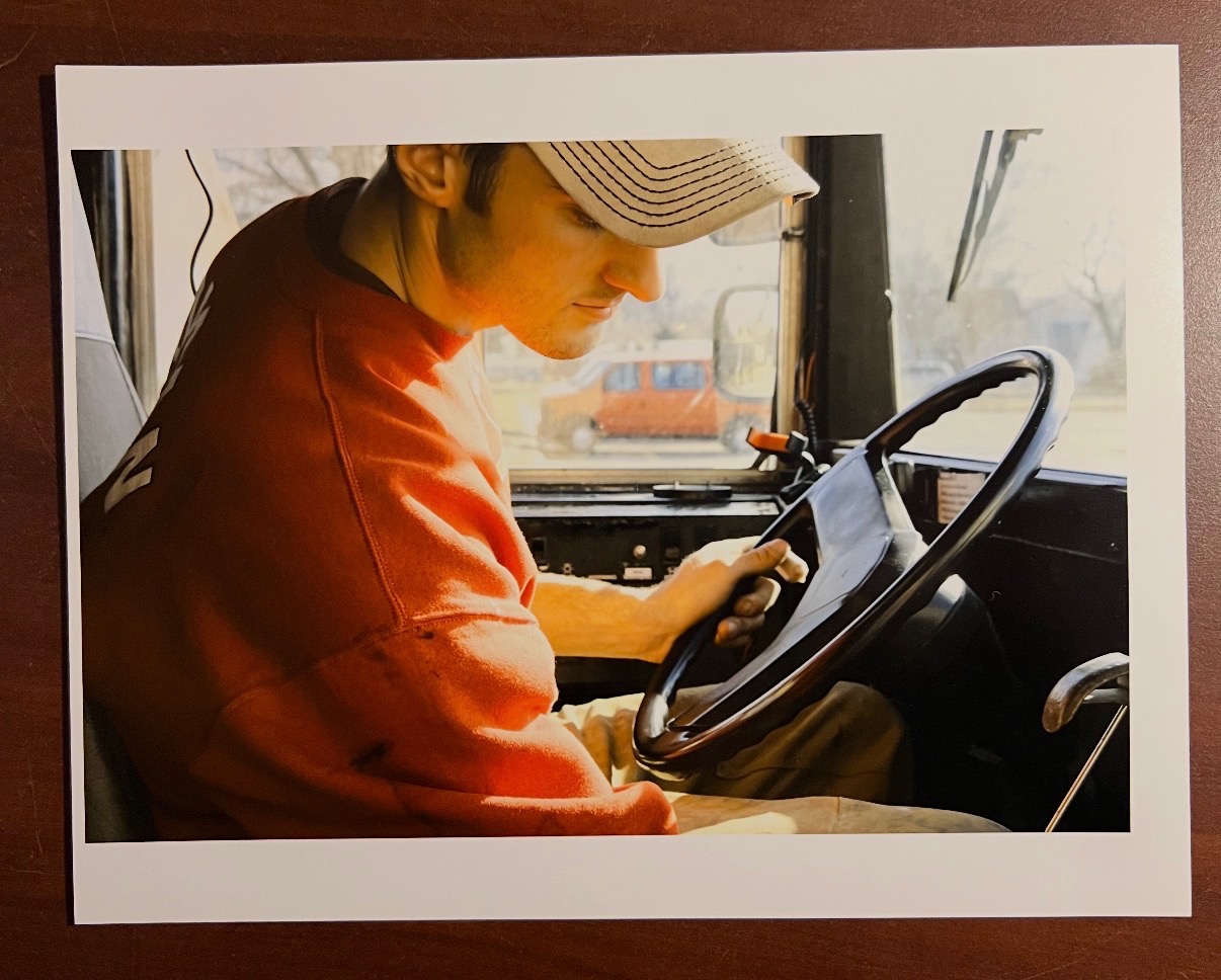 Person wearing a cap and red shirt sitting in a vehicle, looking at a tablet or device, with a steering wheel nearby.