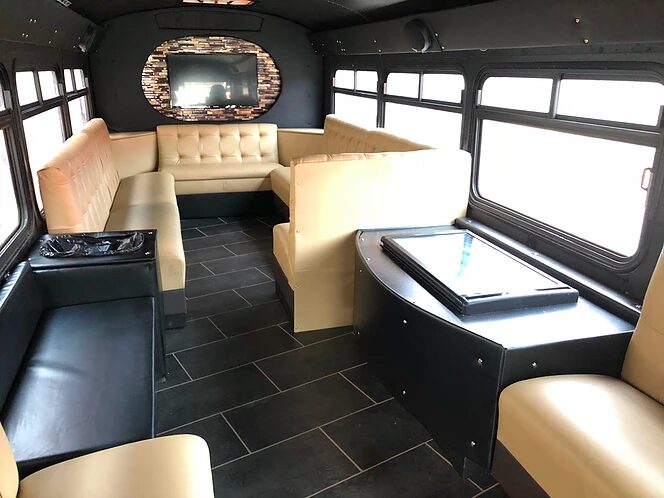Bus interior with beige seating, black floor, windows, and a TV mounted on the wall.