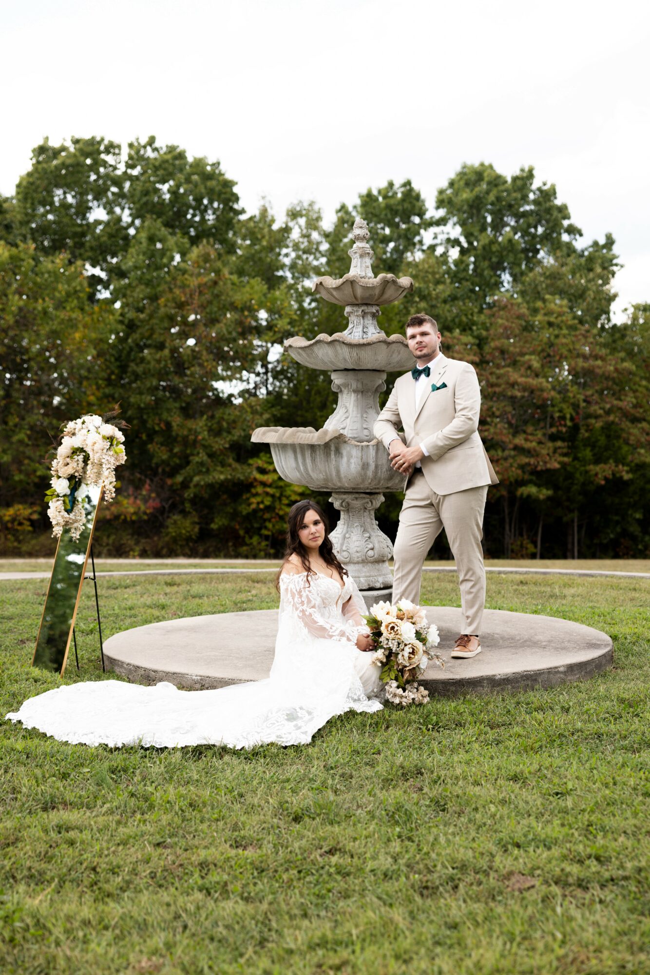 Bride and groom pose in front of a fountain with trees in the background, bride sitting on the grass with flowers, groom standing.